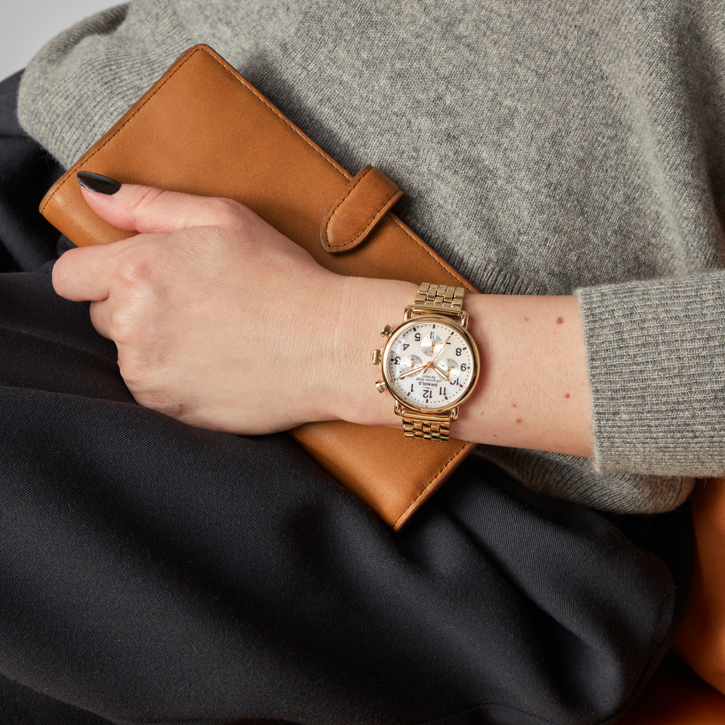 A person wearing a Shinola Runwell Chronograph Watch - MOP Gold Bracelet and a grey sweater holds a brown leather wallet, resting their hand on black fabric.
