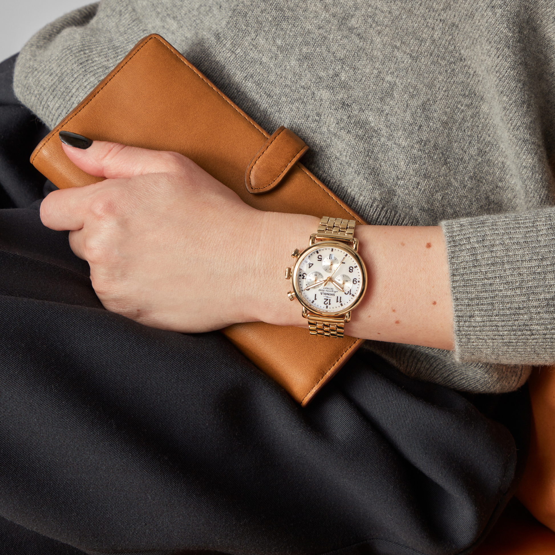 A person wearing a Shinola Runwell Chronograph Watch - MOP Gold Bracelet and a grey sweater holds a brown leather wallet, resting their hand on black fabric.