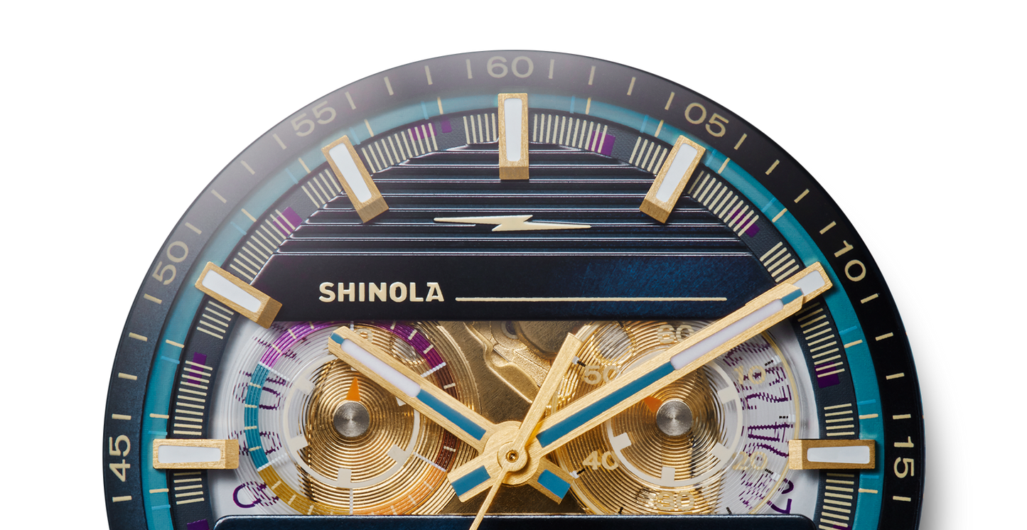 Close-up of a Shinola J Dilla Runwell Chronograph Watch - Midnight Blue face with gold and blue hands, bold hour markers, detailed minute indicators, and visible gears on a dark blue outer ring.