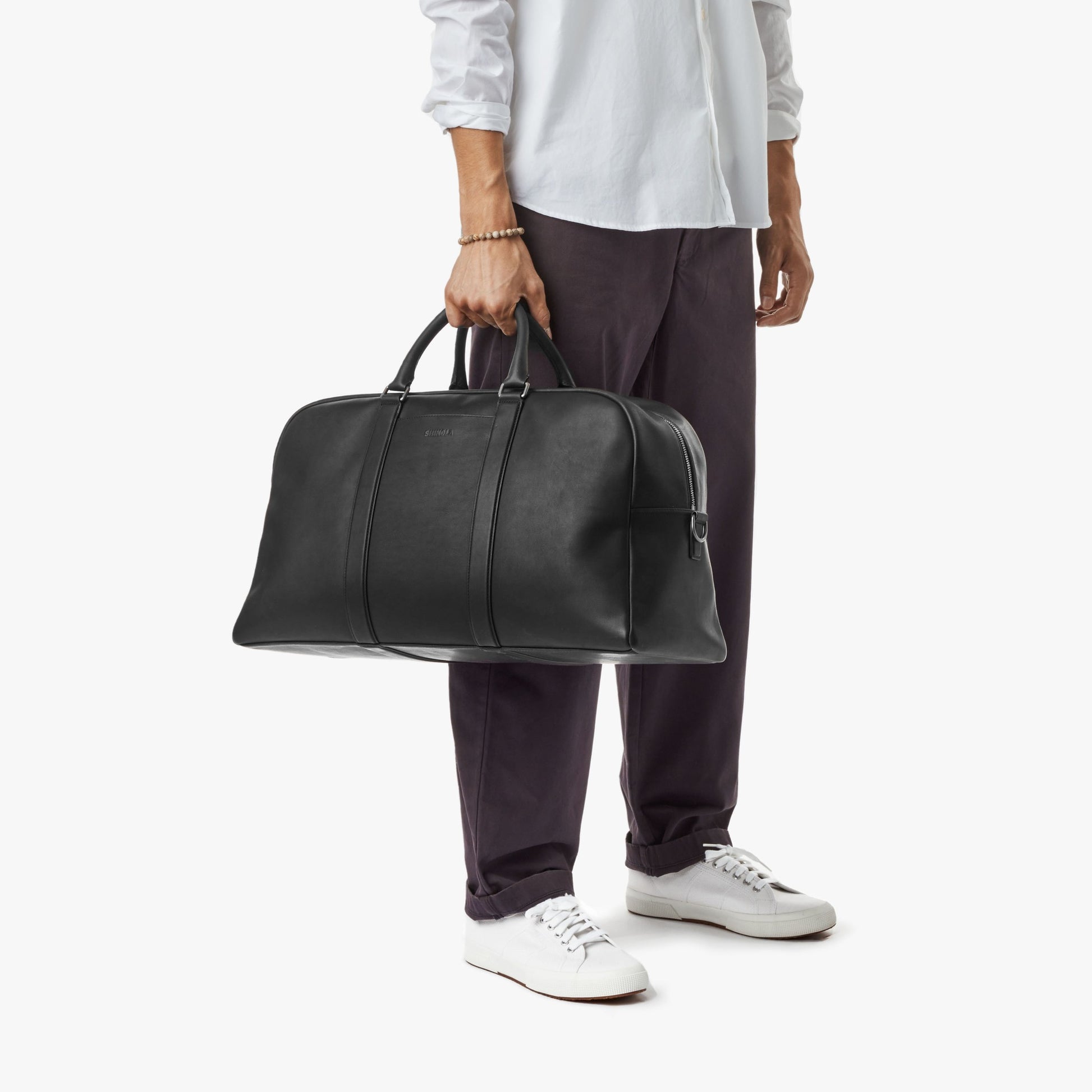 A person dressed in a white shirt, dark pants, and white sneakers holds a Shinola Runwell Duffle Bag - Black by the handles, standing against a plain white background.