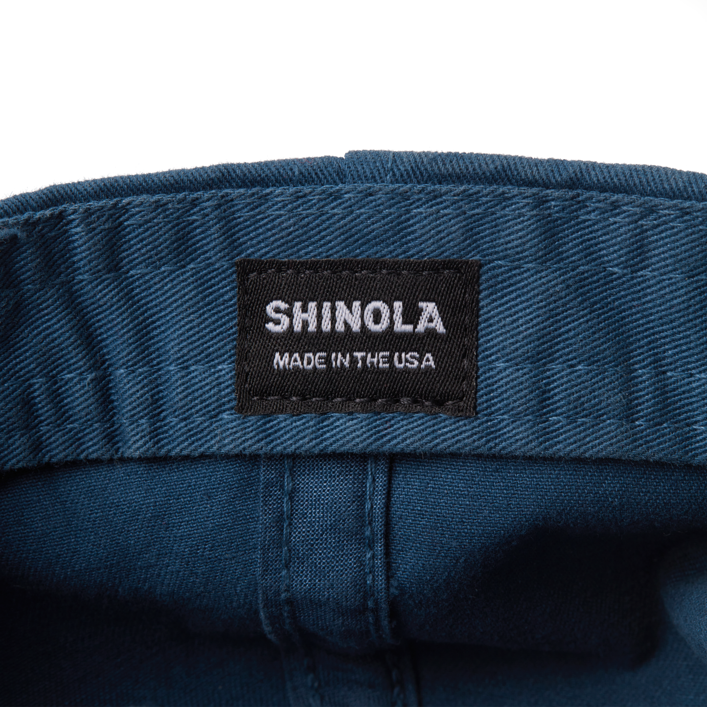 A close-up of a black fabric label sewn onto the blue Cotton Twill 6 Panel Cap by Shinola, displaying the text SHINOLA MADE IN THE USA in white capital letters.