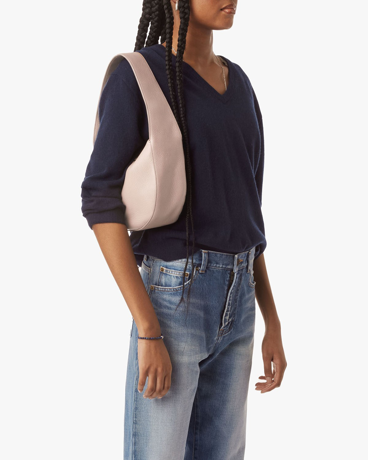 A person with long braids, shown from the shoulders down against a plain background, wears a navy V-neck sweater and blue jeans while carrying the Shinola Small Shoulder Bag in taupe, crafted from natural grain leather.