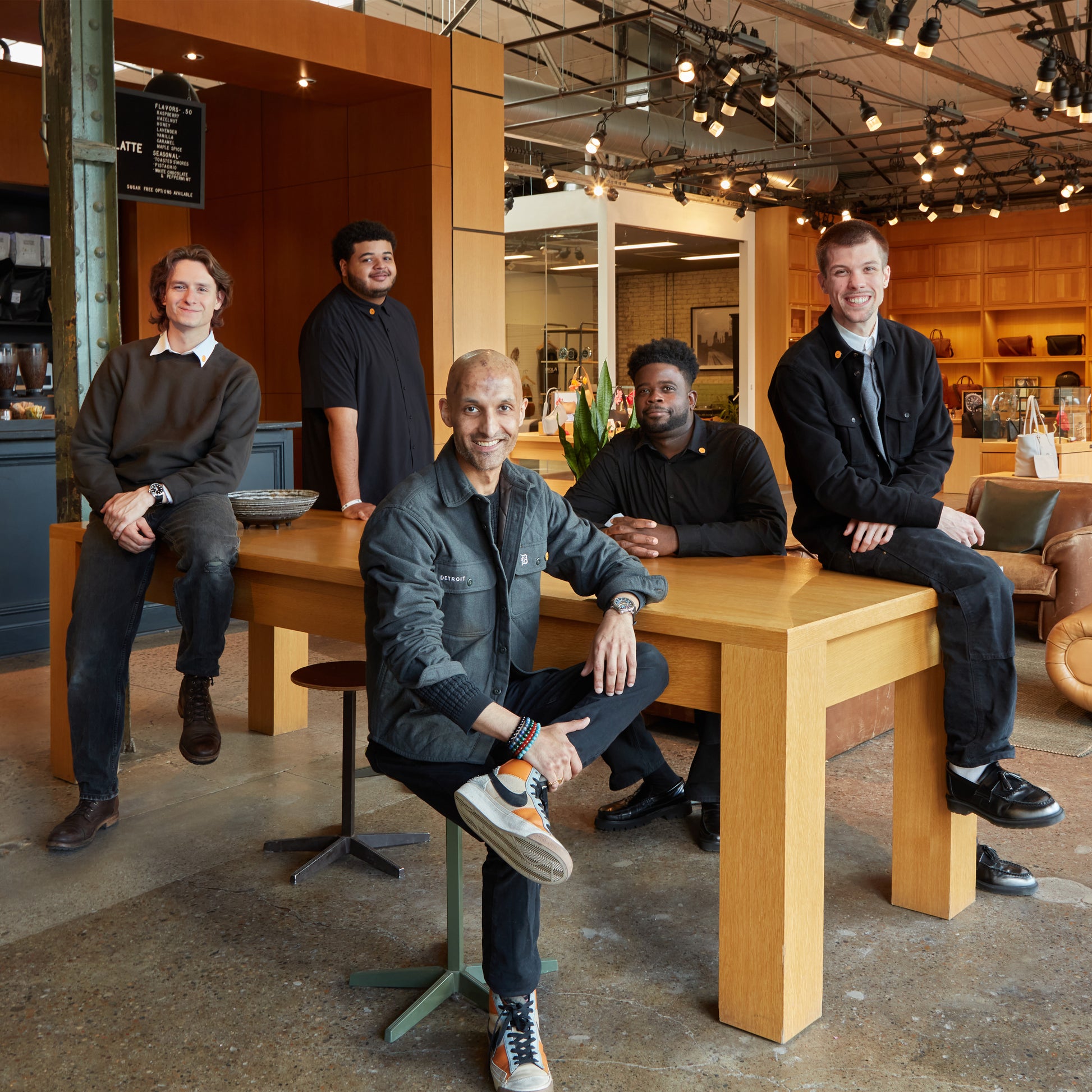 Five people smile together in a modern showroom featuring Shinola watches. Three sit by a large wooden table, two stand behind it, with shelves and spotlights highlighting the space. Experience this with a Shinola Virtual Appointment.