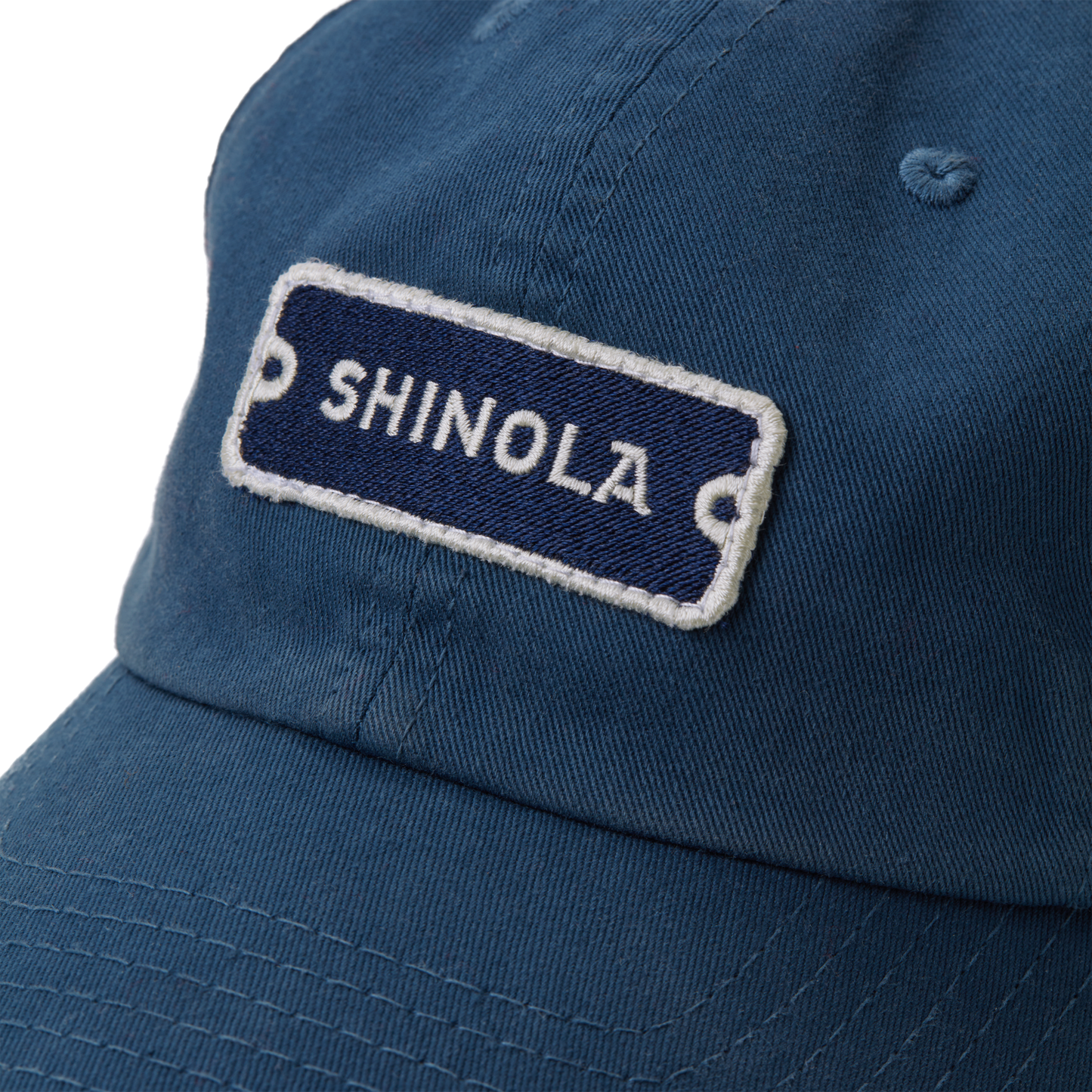 Close-up of the Shinola Cotton Twill 6 Panel Cap in blue, made from garment-dyed cotton twill, featuring a rectangular SHINOLA patch with white letters on a dark blue background and a leather strap closure.