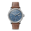 The Shinola Runwell Date Watch features a blue dial with white numerals, date display, silver bezel, and a brown leather strap with visible stitching plus signature Shinola branding.
