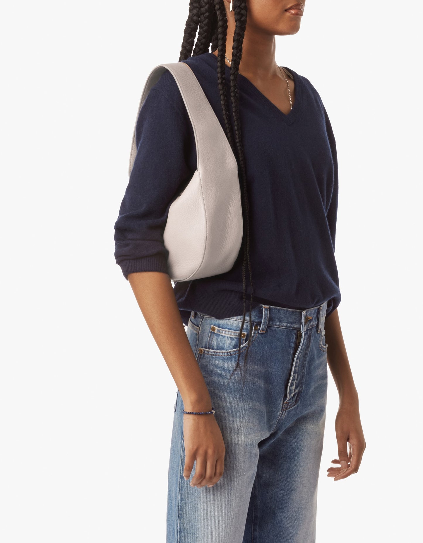 A person with long braids, wearing a navy V-neck sweater and blue jeans, carries the Shinola Small Shoulder Bag in taupe. The bag is crafted from premium leather and the background is plain white.
