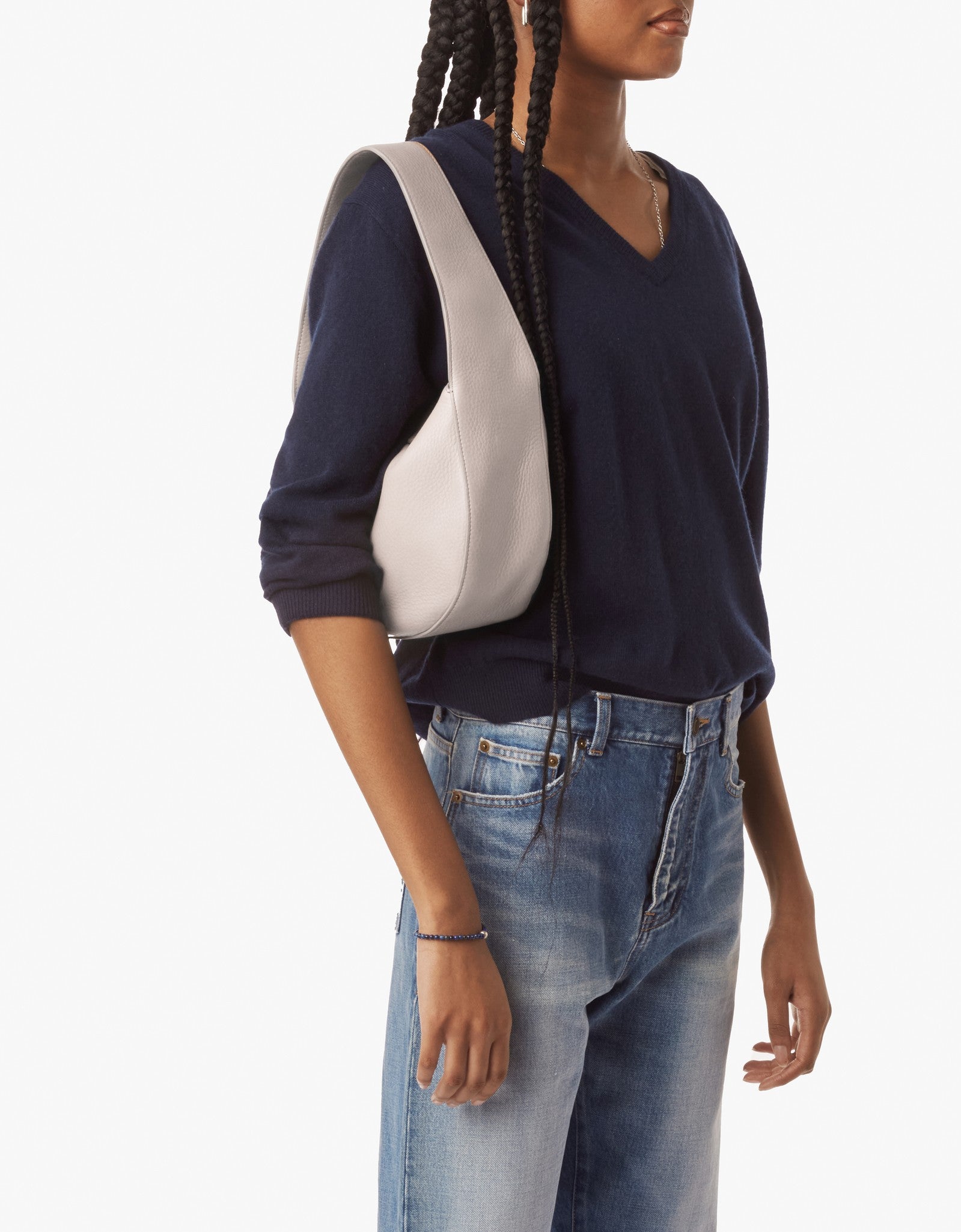 A person with long braids, wearing a navy V-neck sweater and blue jeans, carries the Shinola Small Shoulder Bag in taupe. The bag is crafted from premium leather and the background is plain white.