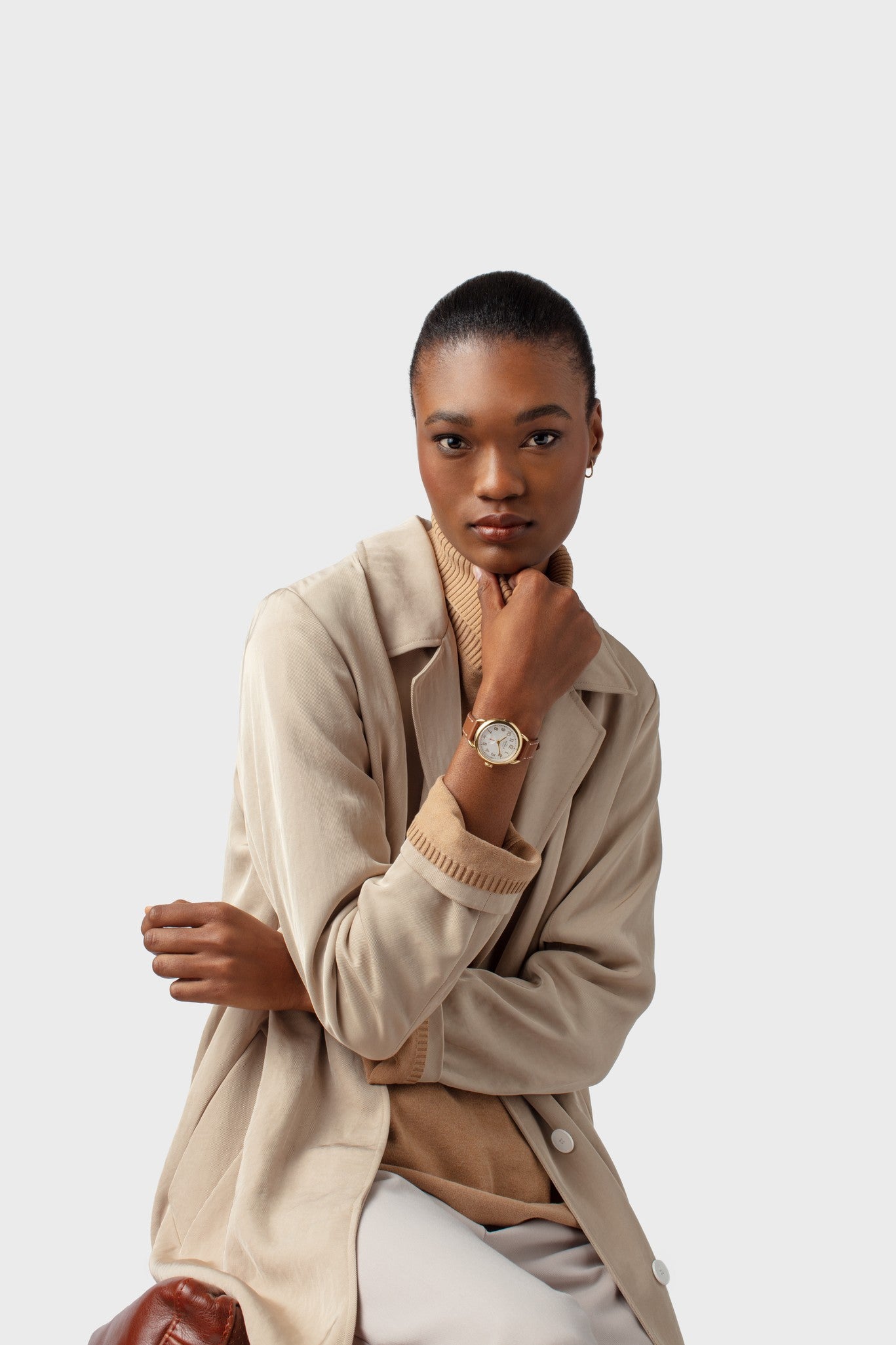 A woman with short hair, in a beige coat and sweater, poses against a light background, showing off the Shinola Runabout Watch - Cream with a leather strap. One arm is folded while her other hand rests by her chin.