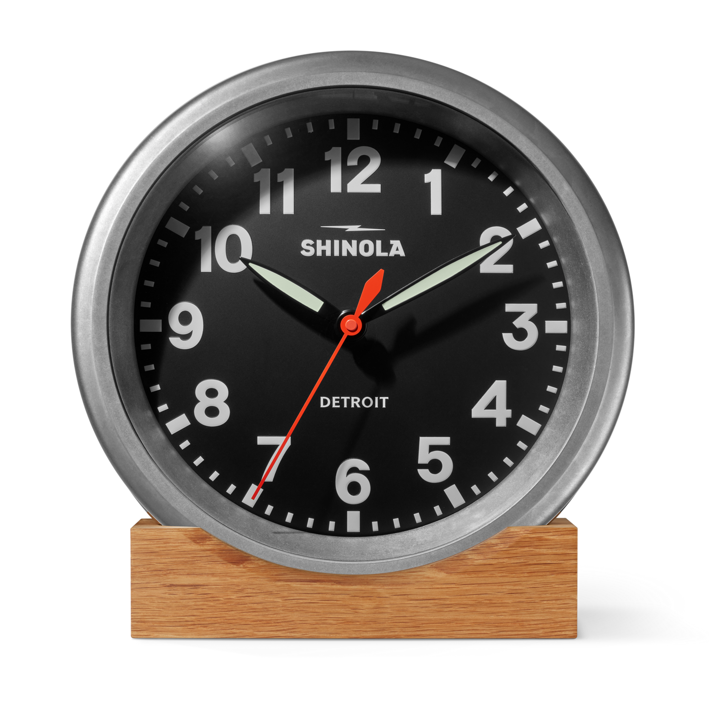 The 14" Runwell Clock by Shinola features a black face, white numbers, and a tumbled zinc frame. It rests on a wooden base, shows 9:10:33, and displays "Shinola" and "Detroit," evoking an industrial clock vibe.