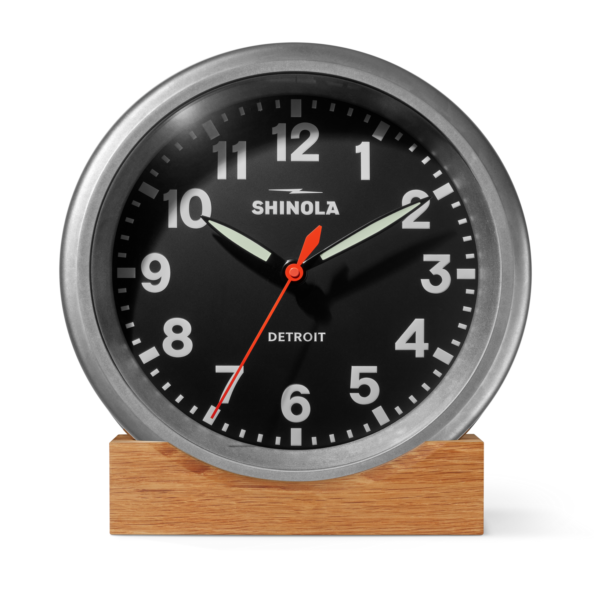 The 14" Runwell Clock by Shinola features a black face, white numbers, and a tumbled zinc frame. It rests on a wooden base, shows 9:10:33, and displays "Shinola" and "Detroit," evoking an industrial clock vibe.
