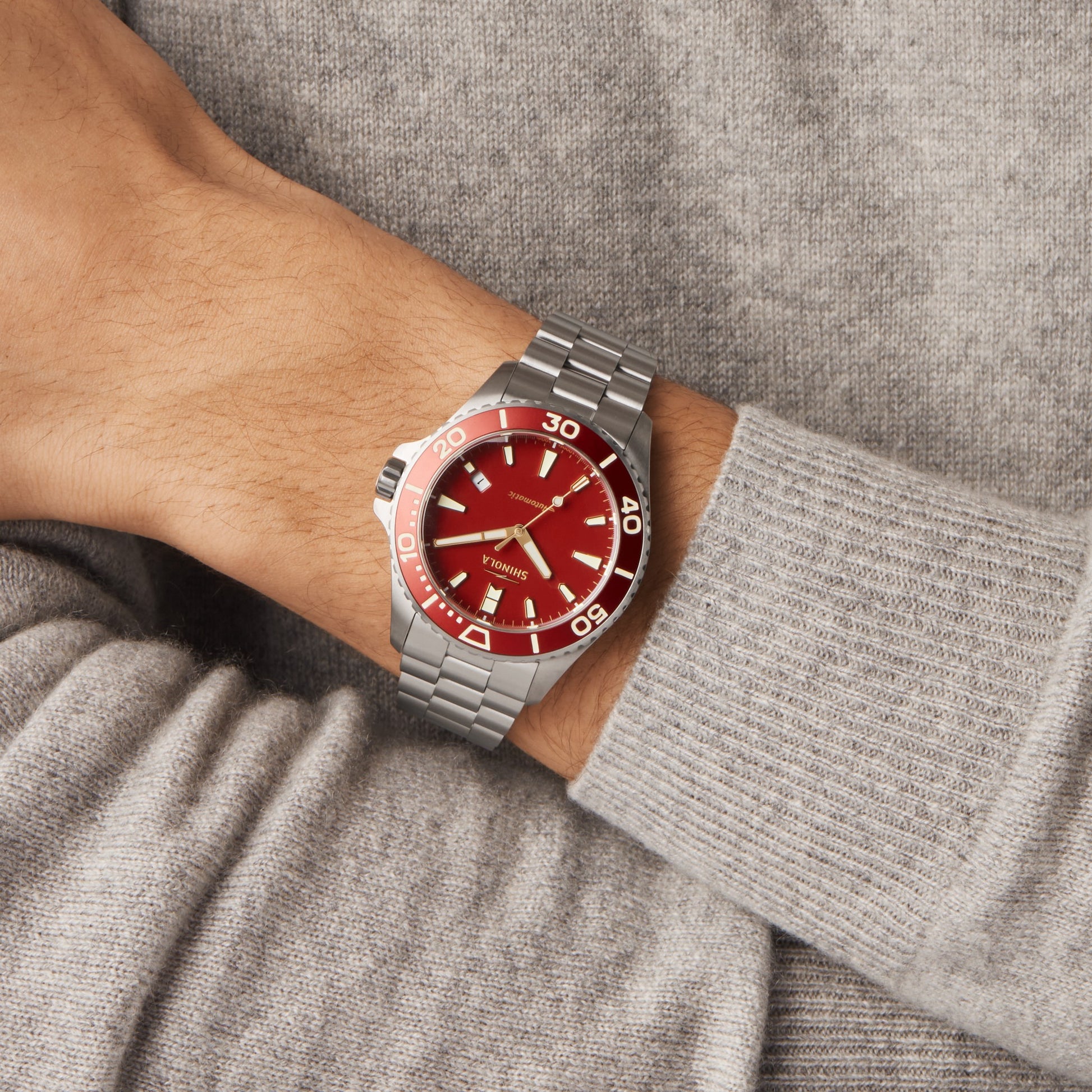 A person in a grey sweater is shown wearing the Shinola Lava Lake Monster Automatic Watch - Cardinal, which features a bold red dial and bezel with white markings.