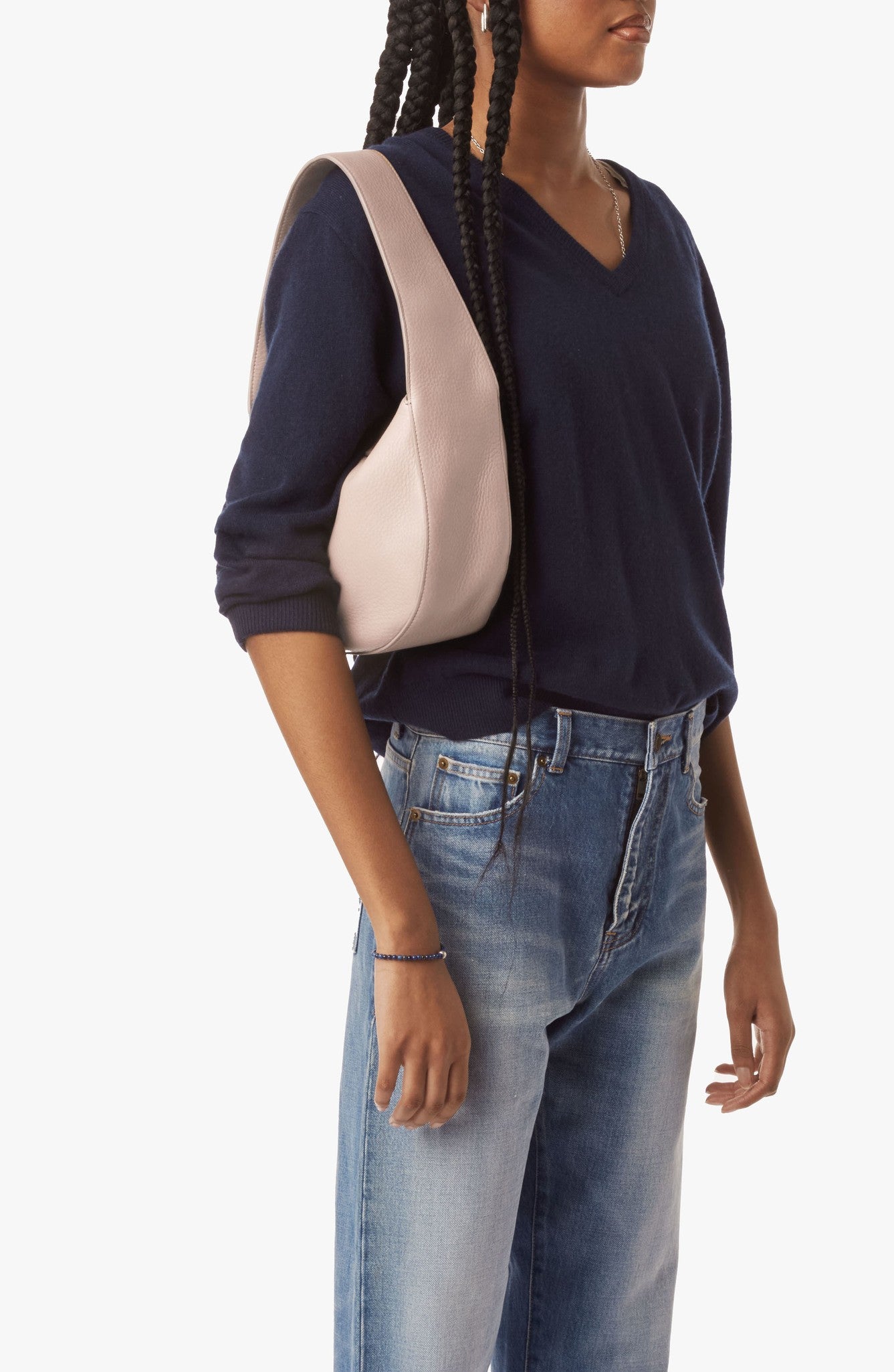 A woman wearing a navy v-neck sweater and light blue jeans stands with the Shinola Small Shoulder Bag in taupe. She has long braided hair, her hand rests by her side, all set against a plain white background.