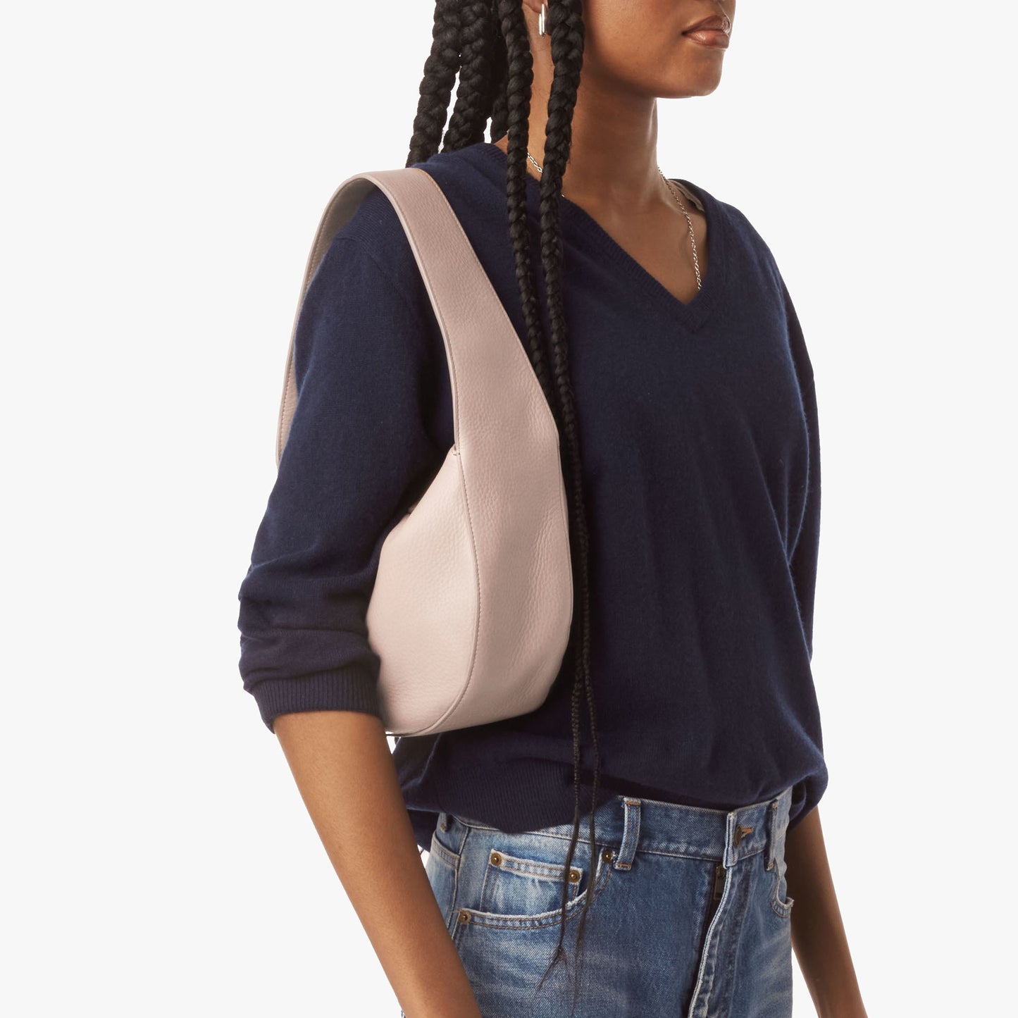 A person with long braids, dressed in a navy V-neck sweater and blue jeans, carries a Shinola Small Shoulder Bag in taupe leather, standing against a plain white background.