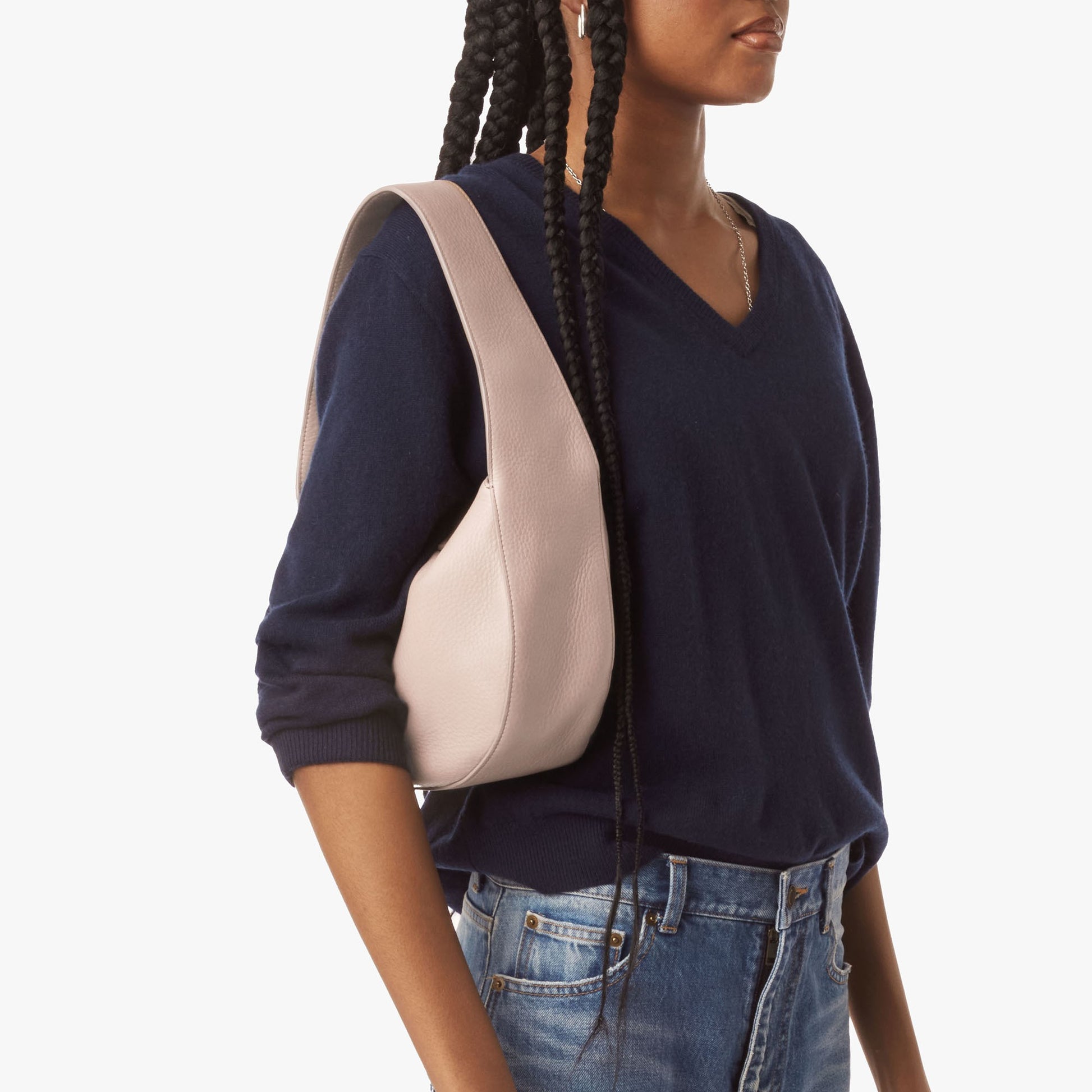 A person with long braids, dressed in a navy V-neck sweater and blue jeans, carries a Shinola Small Shoulder Bag in taupe leather, standing against a plain white background.