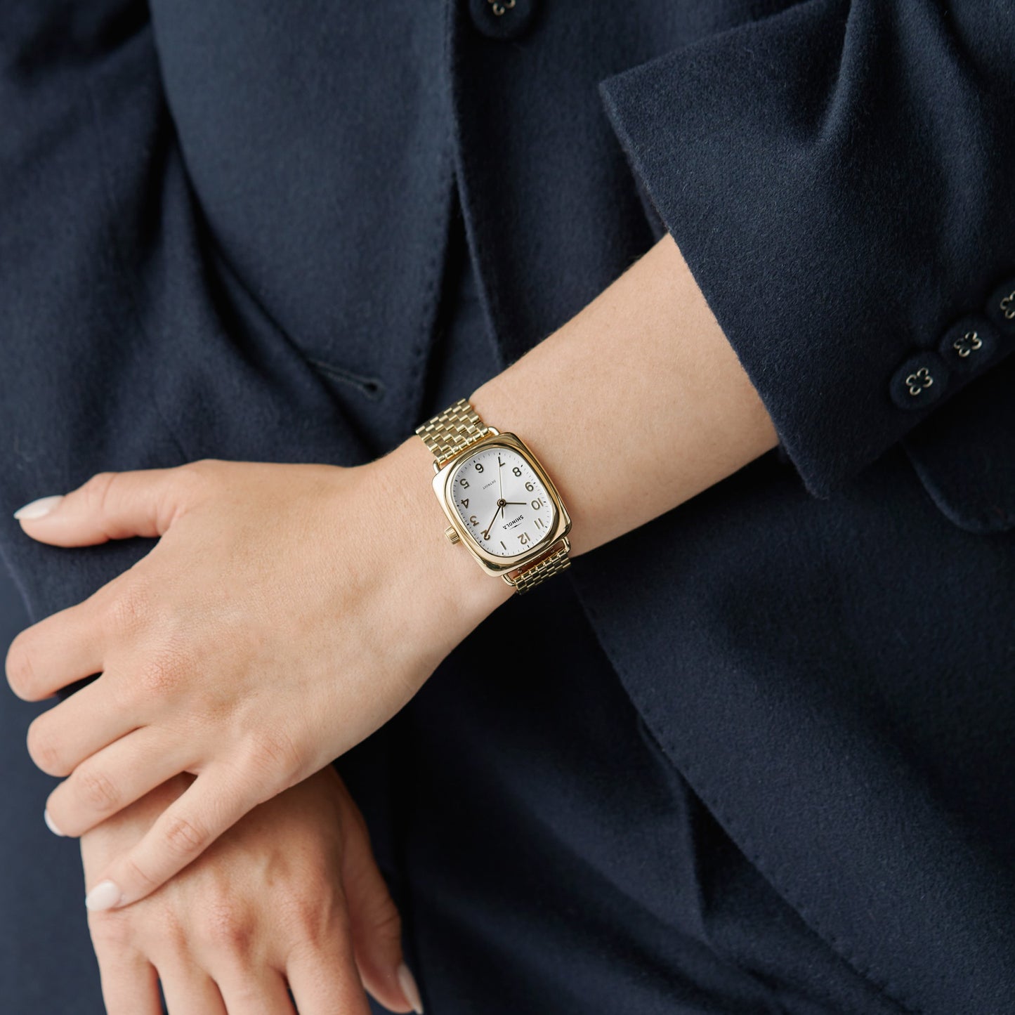 A person in a dark blue blazer crosses their arms, showcasing the Shinola Bixby Watch – Light Silver with a rectangular white face and black numerals.