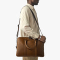 A person in a light beige outfit carries a Shinola Computer Briefcase - Medium Brown with handles and a long strap across their body, standing against a plain white background.