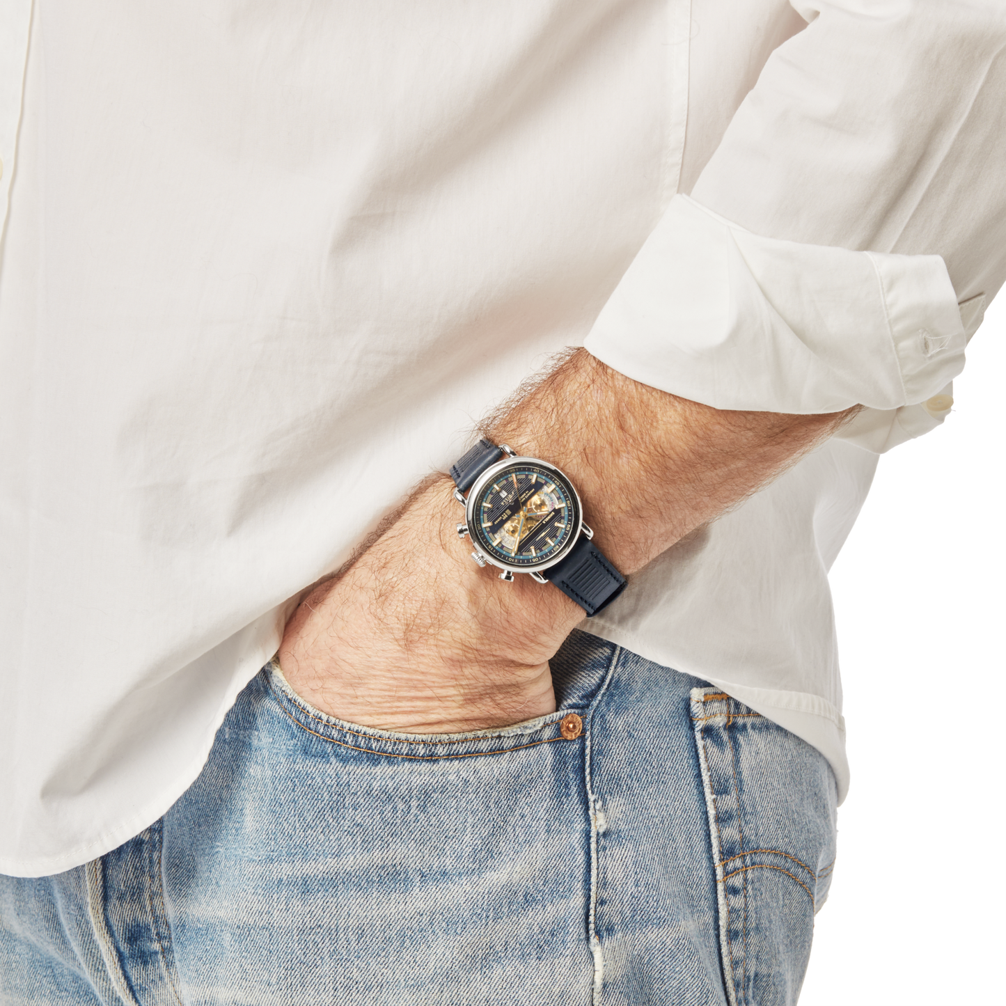 A person in a white shirt and blue jeans has their hand in their pocket, showcasing a Shinola J Dilla Runwell Chronograph Watch - Midnight Blue with a detailed, multi-colored face.