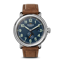 The Shinola Runwell Automatic Watch - Midnight Blue features a brown leather strap, round blue dial, white and yellow numerals, date at 3 o’clock, and a small seconds subdial above 6—exemplifying quality craftsmanship.