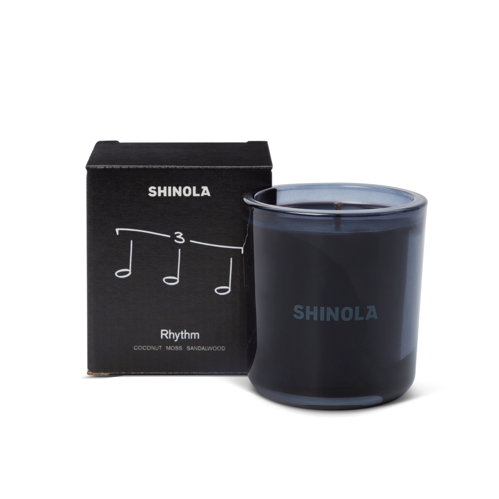 The Shinola Rhythm 8 oz Scented Candle sits next to its box decorated with musical notes and "Rhythm," inspired by Detroit nightlife. Scented with coconut, moss, and sandalwood, the candle comes in a recycled glass vessel.