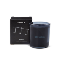 The Shinola Rhythm 8 oz Scented Candle sits next to its box decorated with musical notes and "Rhythm," inspired by Detroit nightlife. Scented with coconut, moss, and sandalwood, the candle comes in a recycled glass vessel.