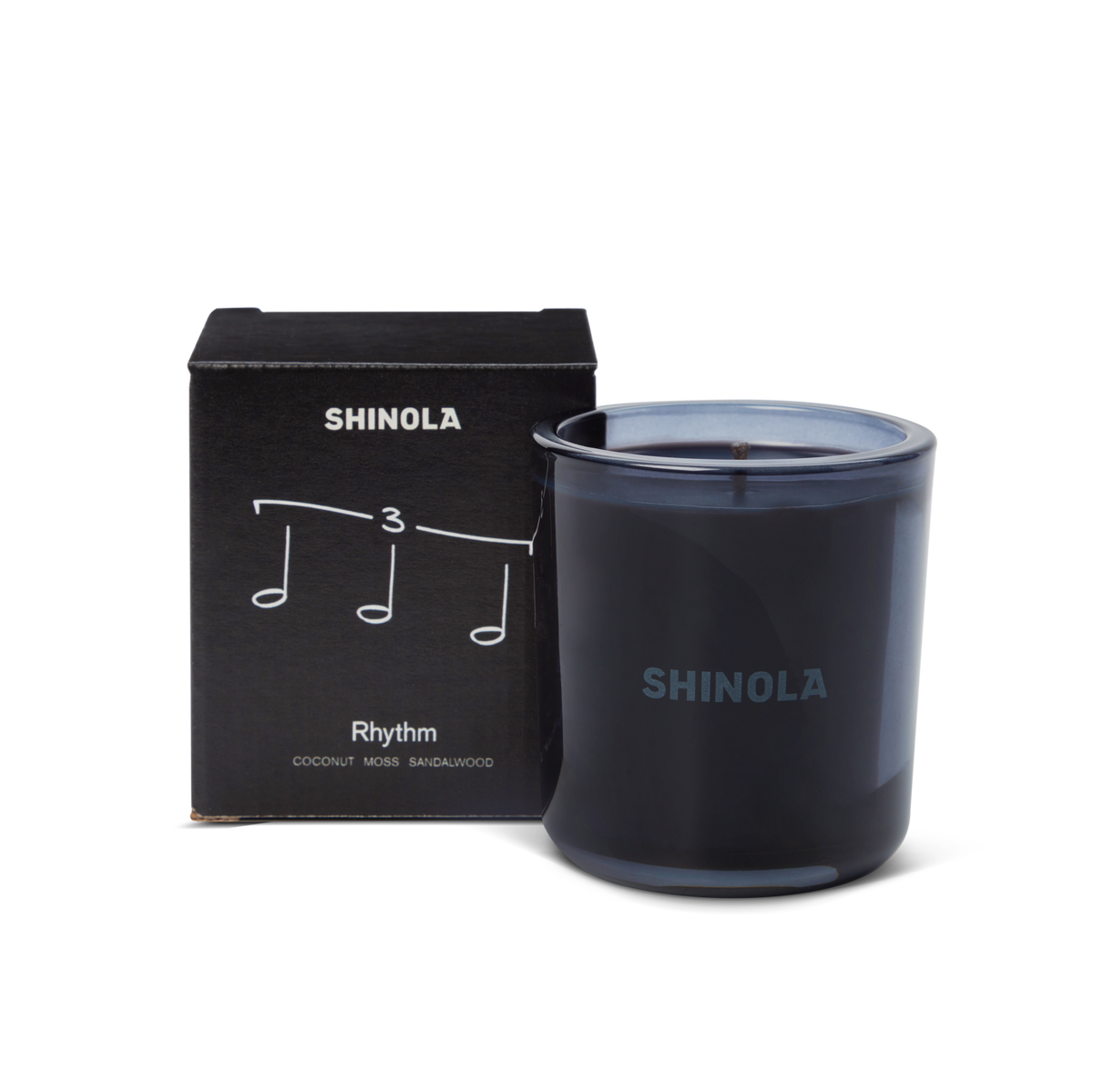 The Shinola Rhythm 8 oz Scented Candle sits next to its box decorated with musical notes and "Rhythm," inspired by Detroit nightlife. Scented with coconut, moss, and sandalwood, the candle comes in a recycled glass vessel.