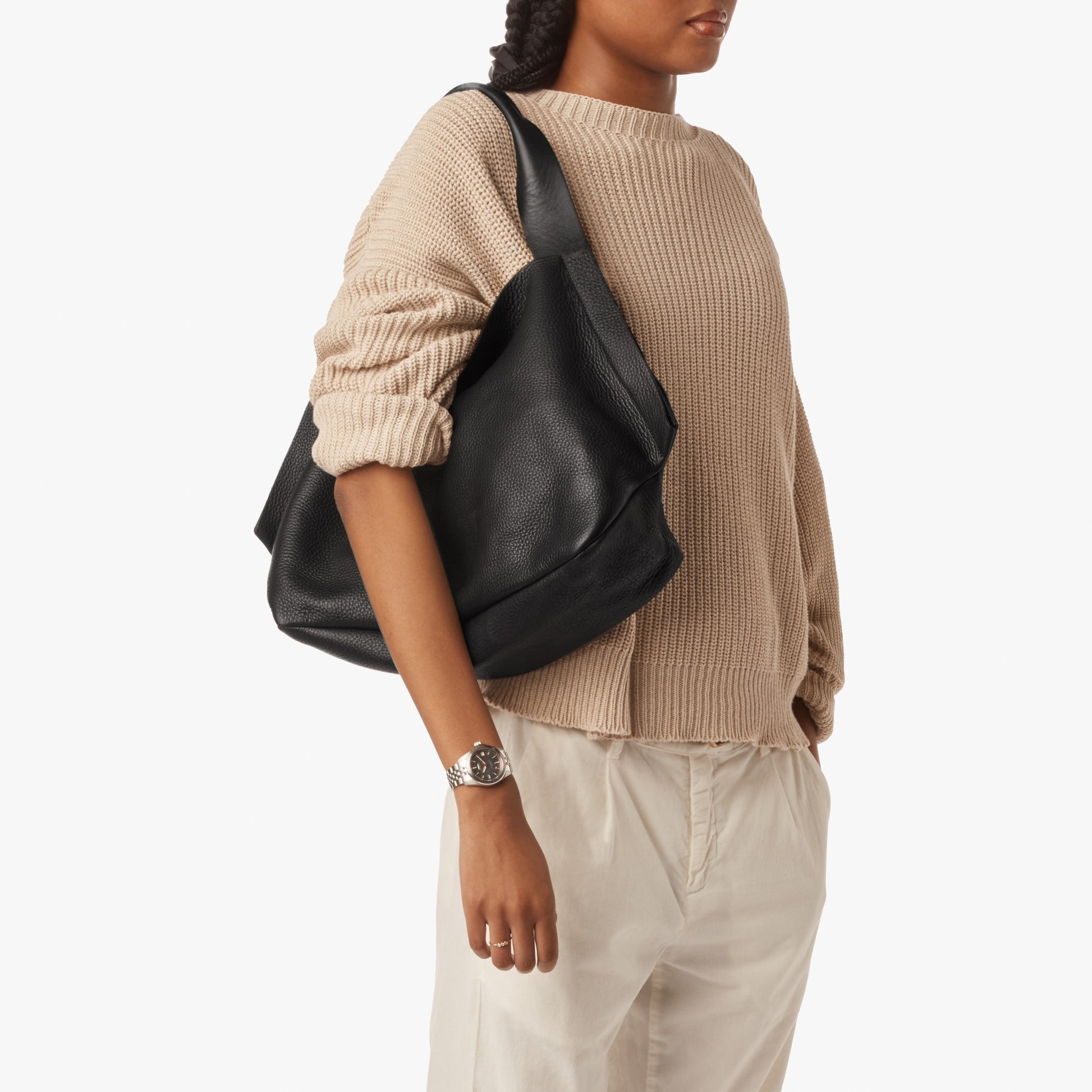 A person in a beige knit sweater and light pants carries the Shinola Derby Shoulder Bag - Black, featuring a leather card pocket and large open compartment. They wear a wristwatch on their left wrist against a plain white background.