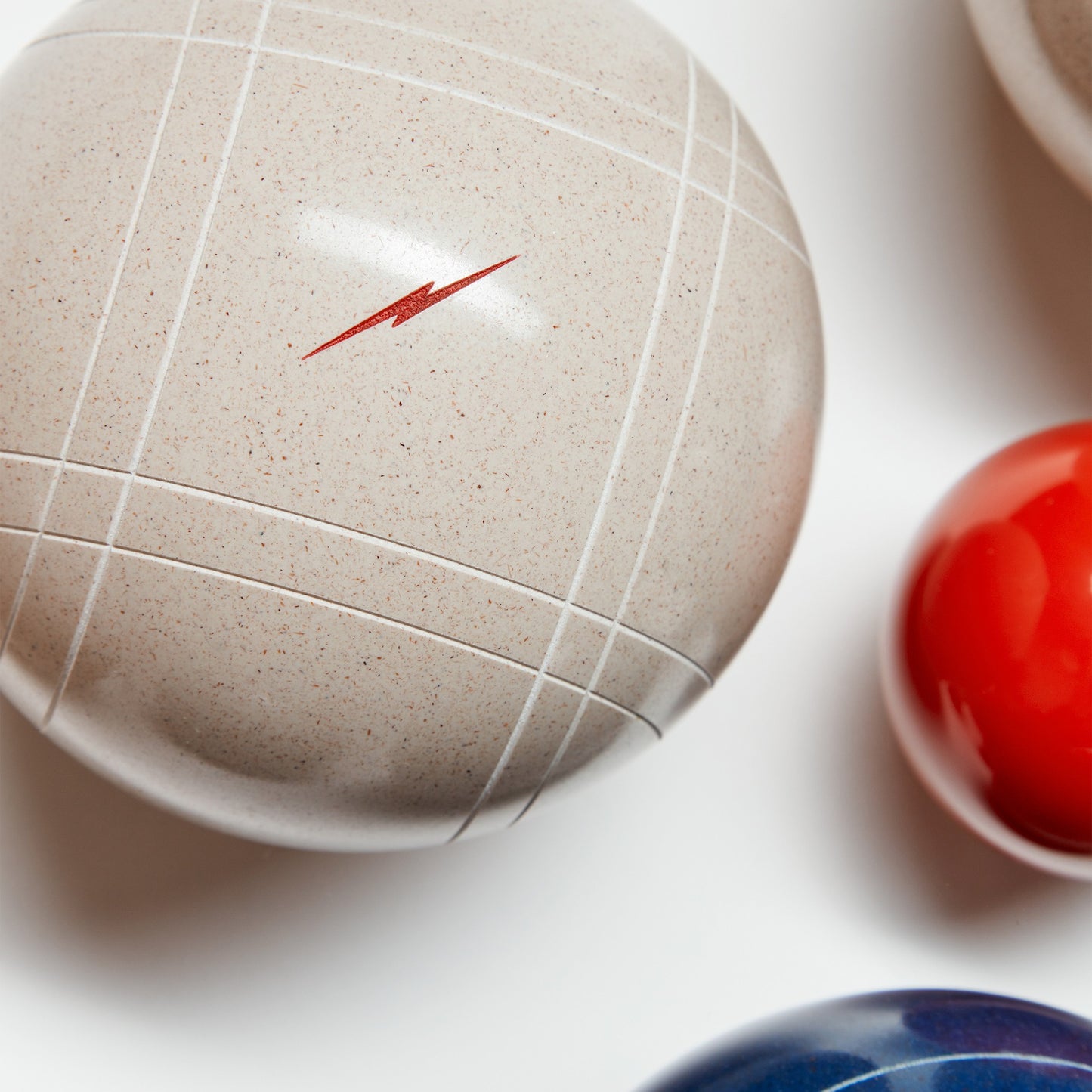 A close-up of three glossy balls from the Shinola + St. Pierre Bocce Ball Set by Shinola: a large beige ball with white lines and a red lightning bolt, plus partial views of red and blue balls, all resting on a white surface.