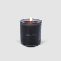 A Shinola Rhythm 8 oz Scented Candle glows warmly in a dark recycled glass vessel, “SHINOLA” printed on the front, set against a plain light gray background.