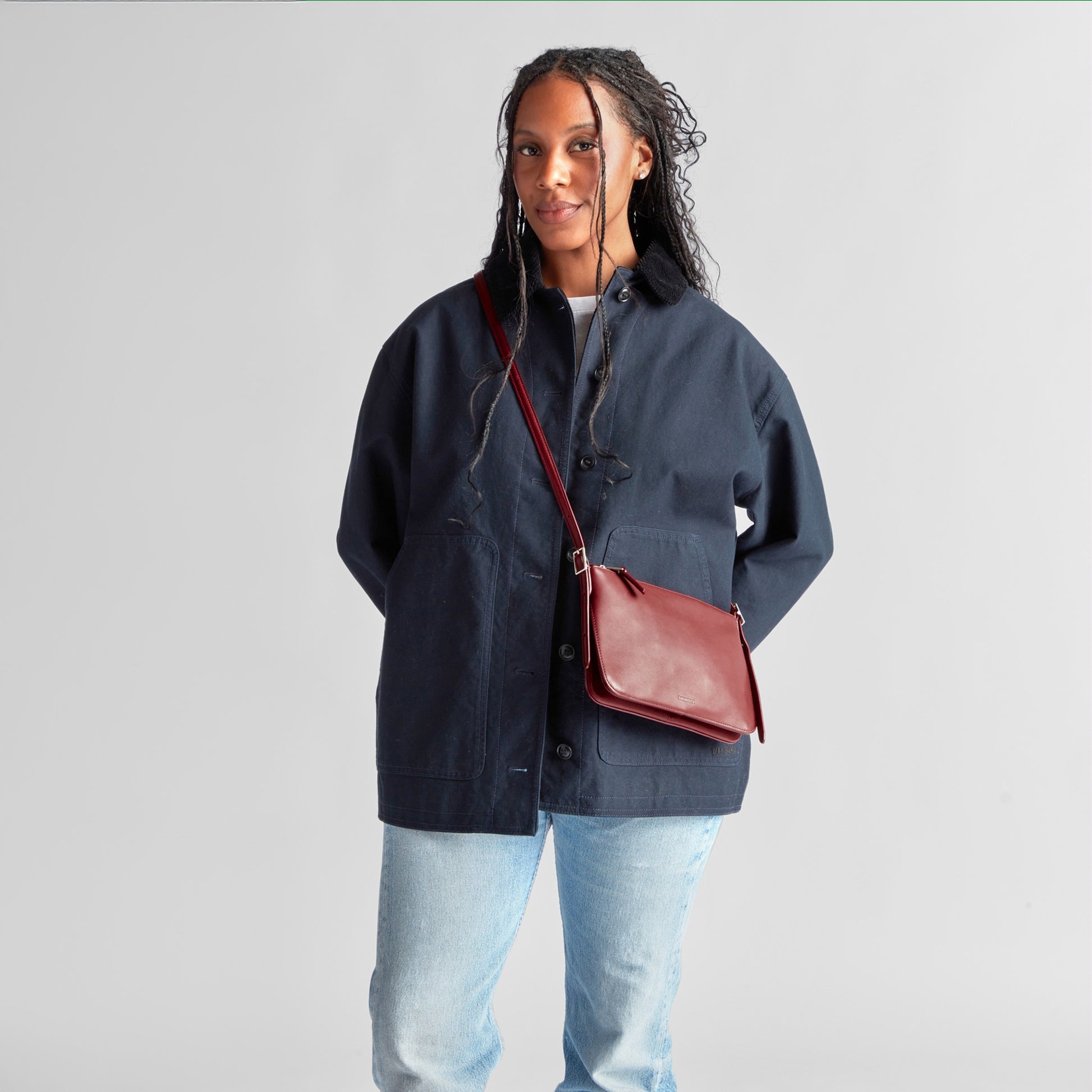 A person stands against a plain background wearing a navy jacket, light blue jeans, and the Shinola Small Bixby Crossbody - Ruby, with their hands partially in their pockets—an ideal everyday accessory.