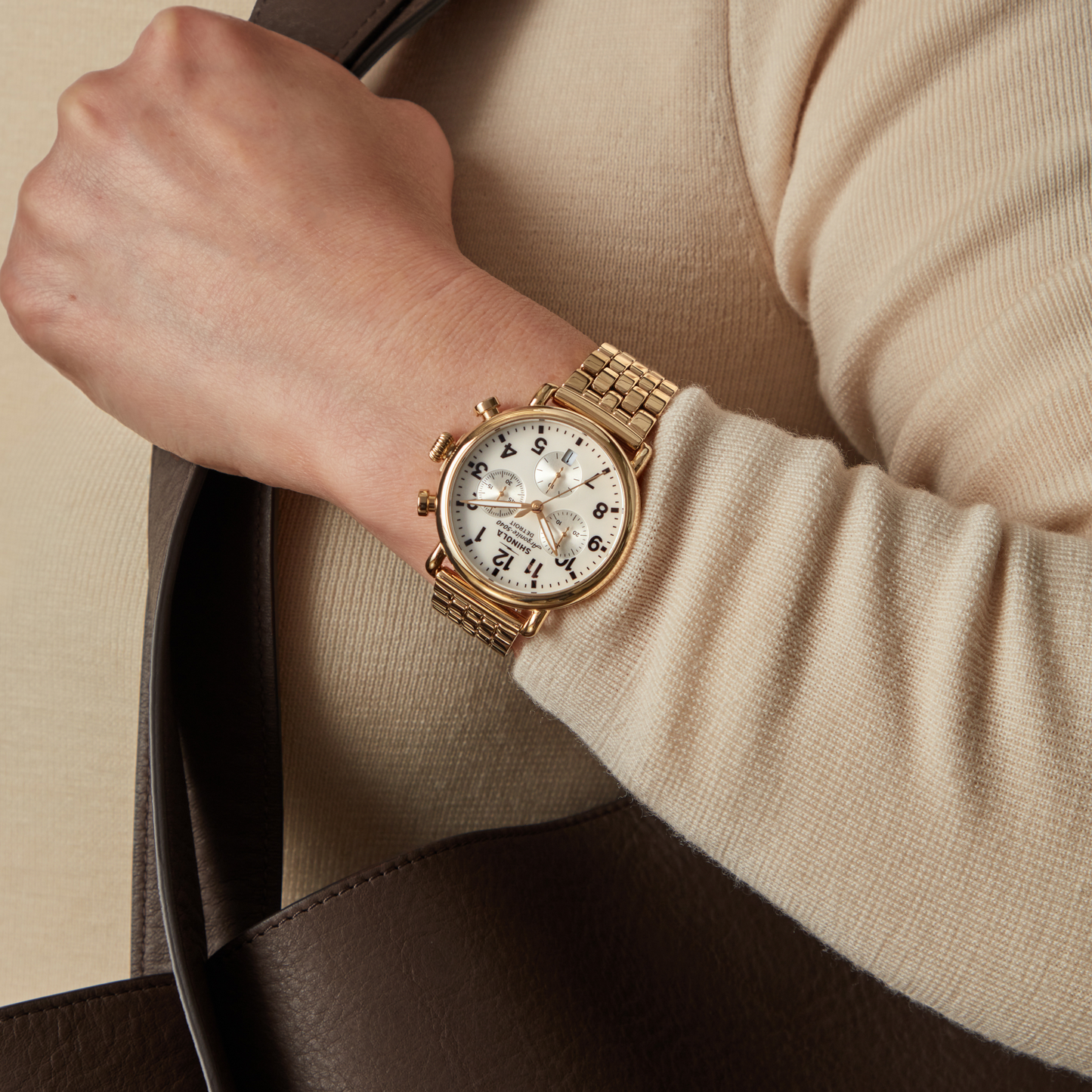A person in a beige long-sleeve top with a brown bag is wearing a Shinola Runwell Chronograph Watch - MOP Gold Bracelet on their wrist.