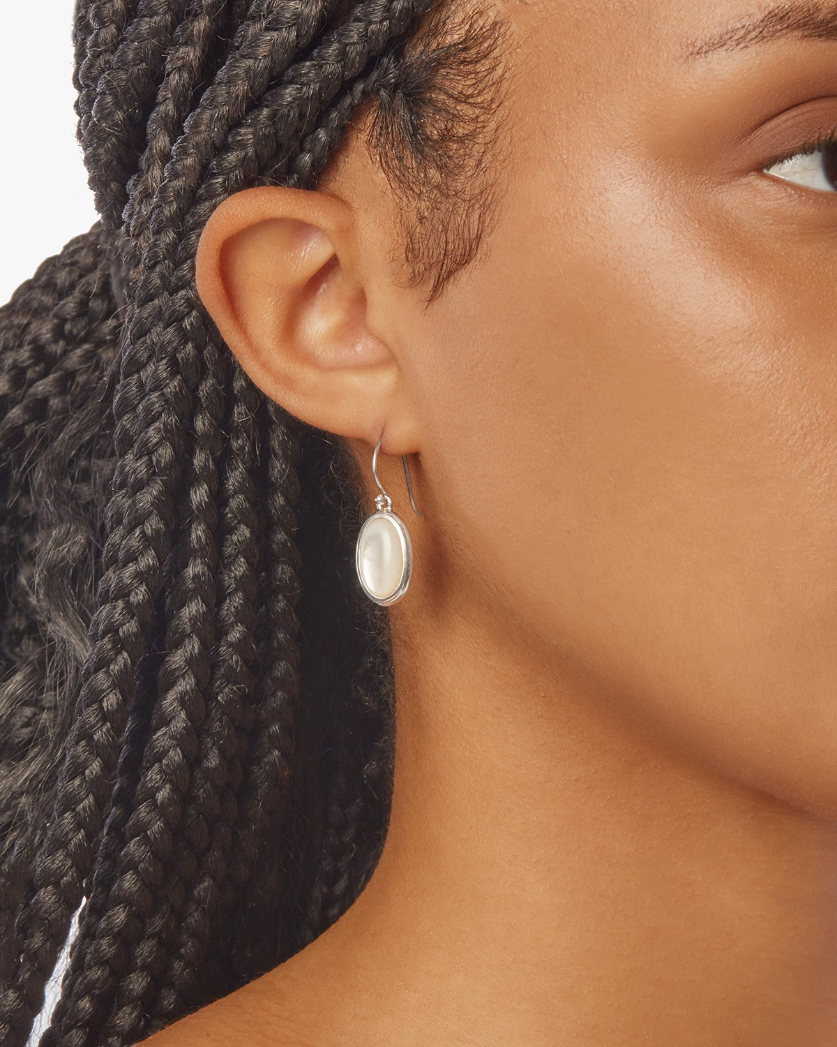 Close-up of a person's ear with braided hair, wearing the Shinola Heirloom Mother Of Pearl Drop Earrings – Sterling Silver. The oval white stone and silver hook stand out against a plain white background.