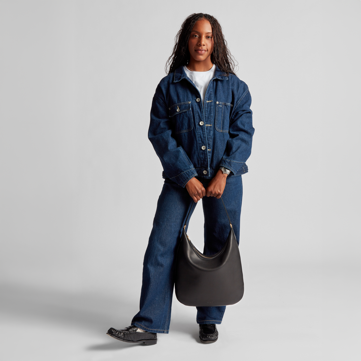 A person stands facing forward in a denim jacket, jeans, and white shirt, holding the Shinola Bixby Shoulder Bag – Black with both hands. Its black leather and accordion-style design provide elegance against a plain light backdrop.