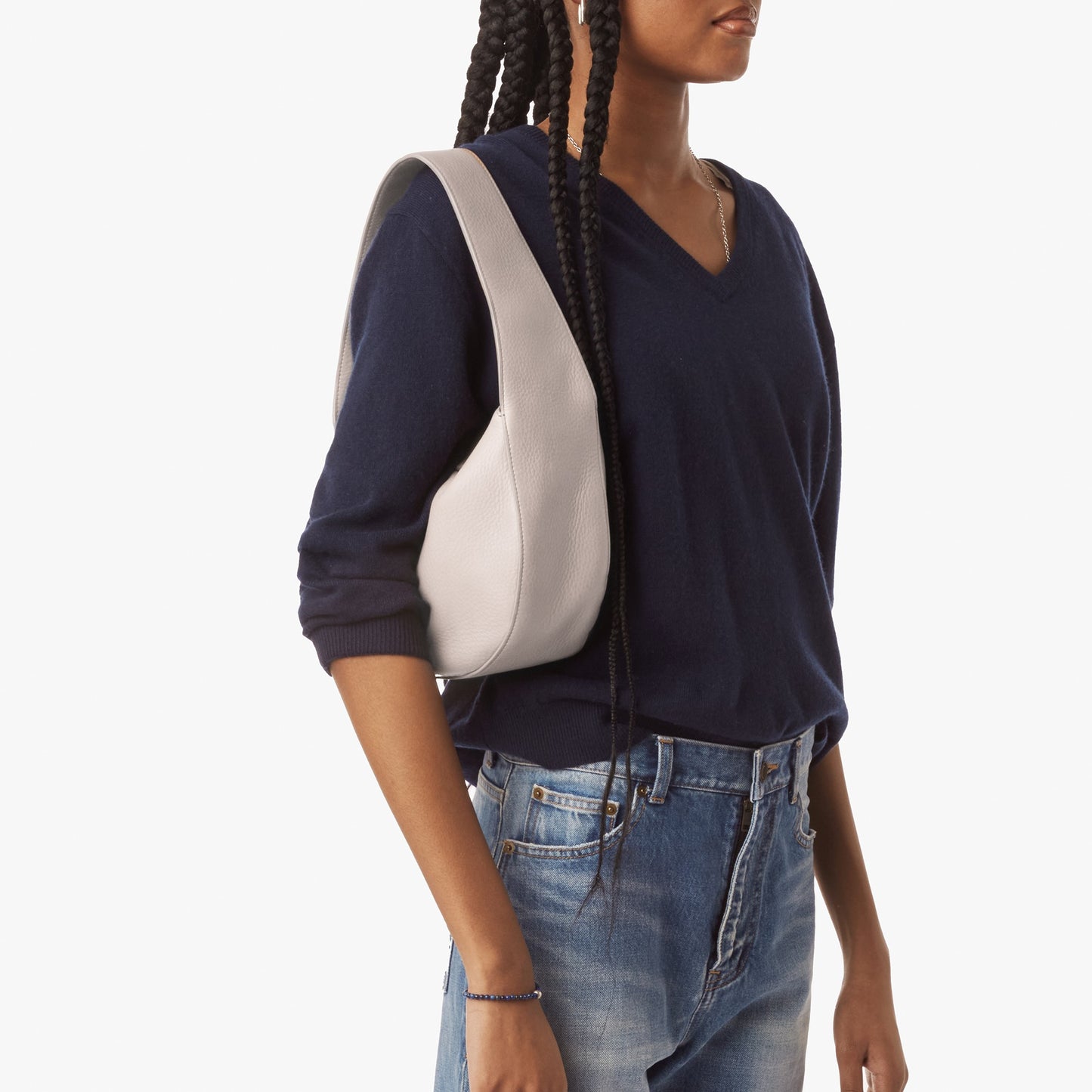 A person with long braids, shown from shoulders to hips against a plain white background, wears a navy V-neck sweater and blue jeans while carrying the Shinola Small Shoulder Bag in Taupe crafted from natural grain leather.