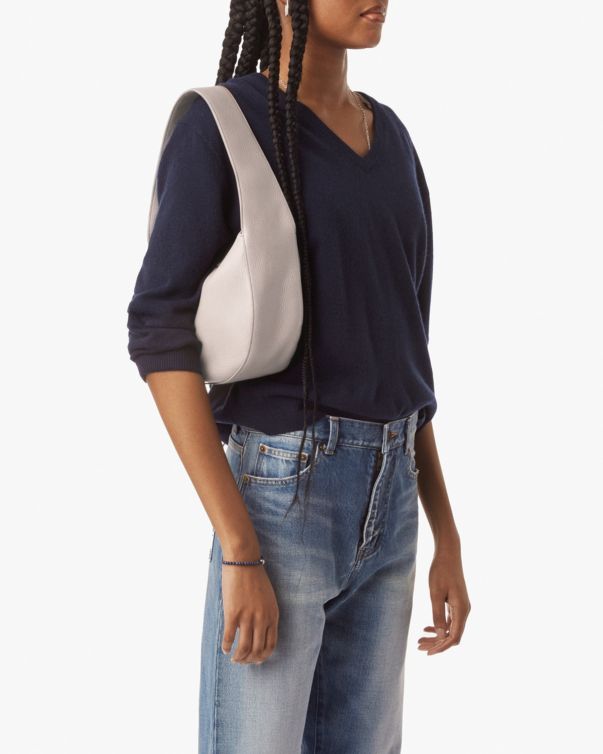 A person with long braids, dressed in a navy V-neck sweater and blue jeans, carries the Shinola Small Shoulder Bag in taupe natural grain leather while standing against a plain white background.