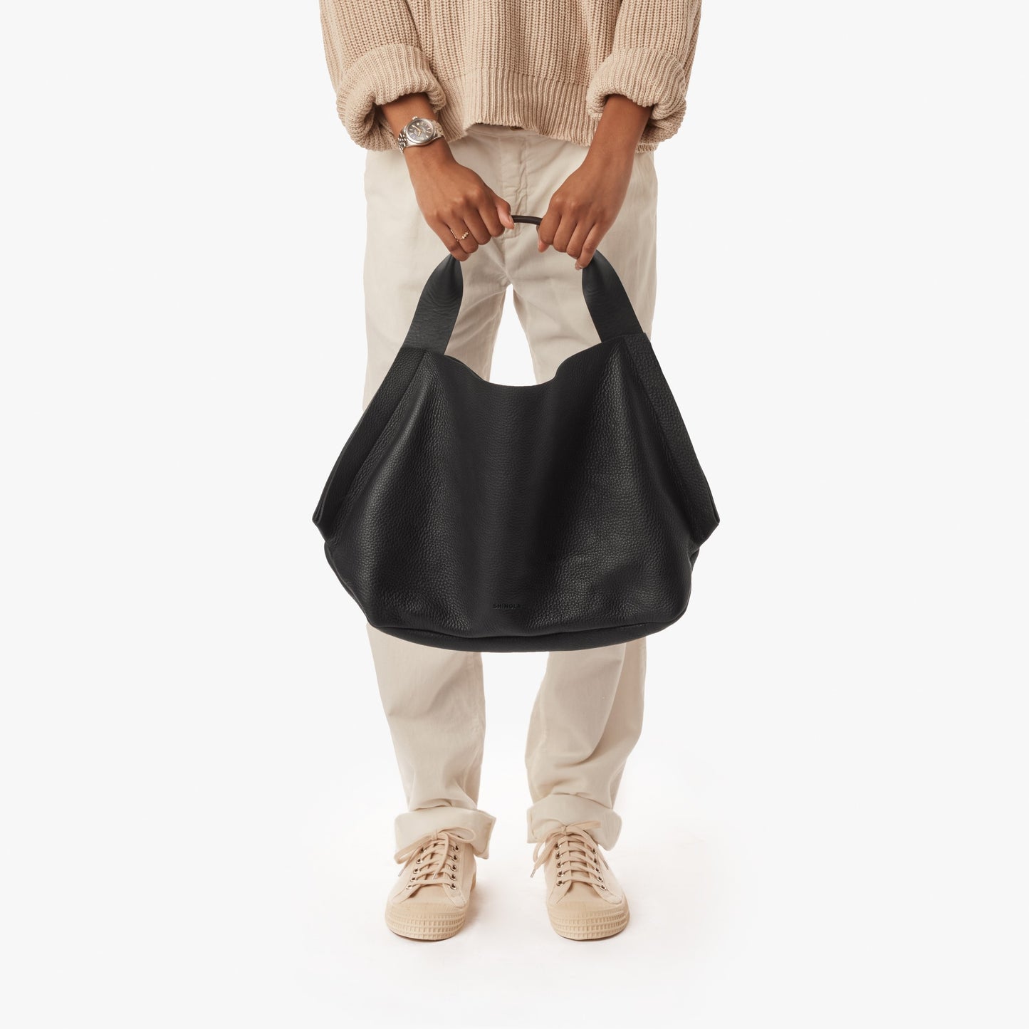 A person in a beige sweater, light pants, and beige sneakers holds the Shinola Derby Shoulder Bag - Black by its handles, showcasing its large open compartment against a plain white background. The image is cropped above the shoulders.