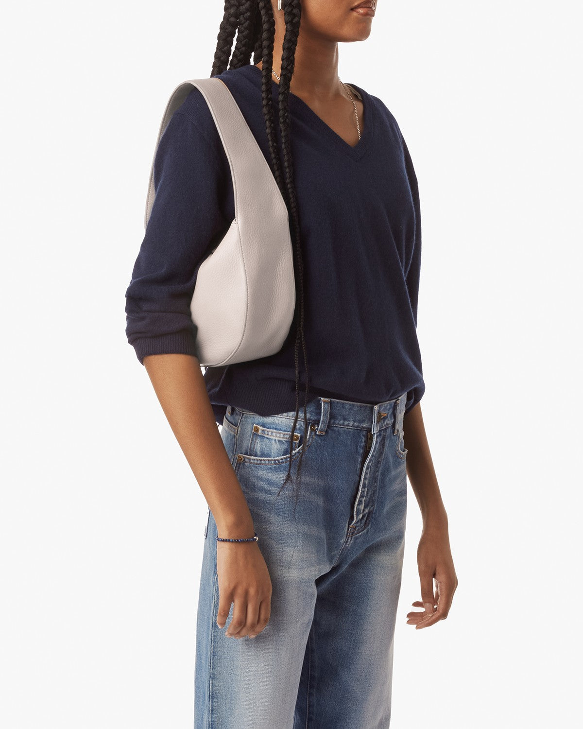 A person in a navy V-neck sweater and blue jeans holds the Shinola Small Shoulder Bag - Taupe. They have long braids and their left arm rests by their side, against a plain white background.
