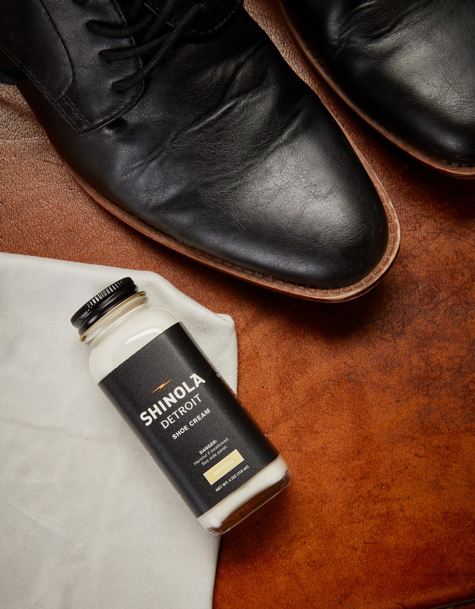 A jar of Shinola Shoe Cream - Neutral rests on a cloth beside polished black leather shoes atop a brown surface.