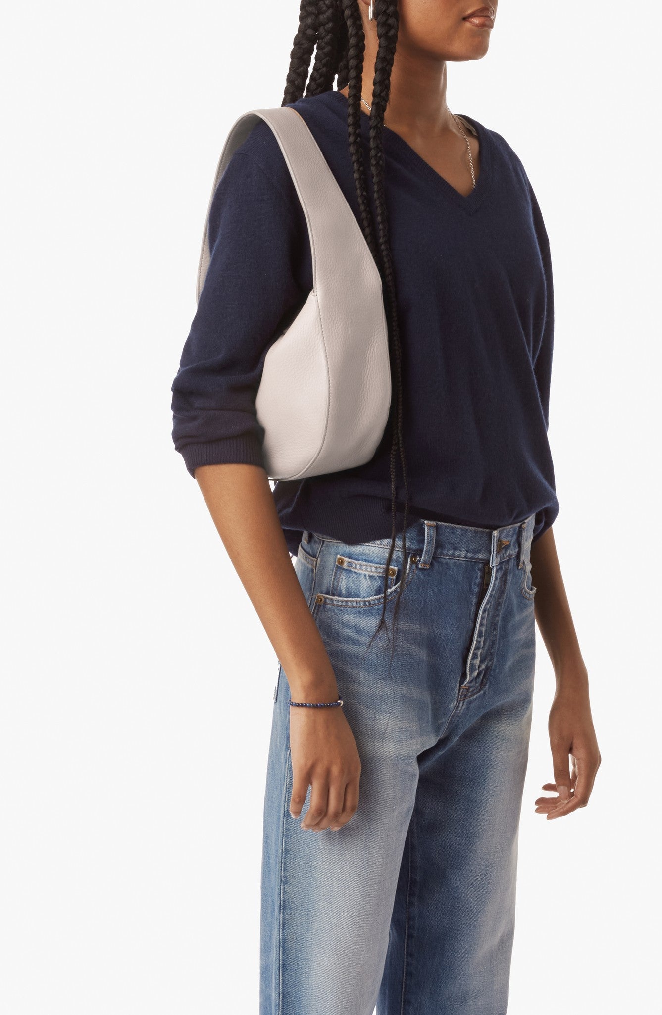 A woman with long braids wears a navy V-neck sweater, blue jeans, and carries the Shinola Small Shoulder Bag in Taupe, crafted from premium leather, against a plain white background.
