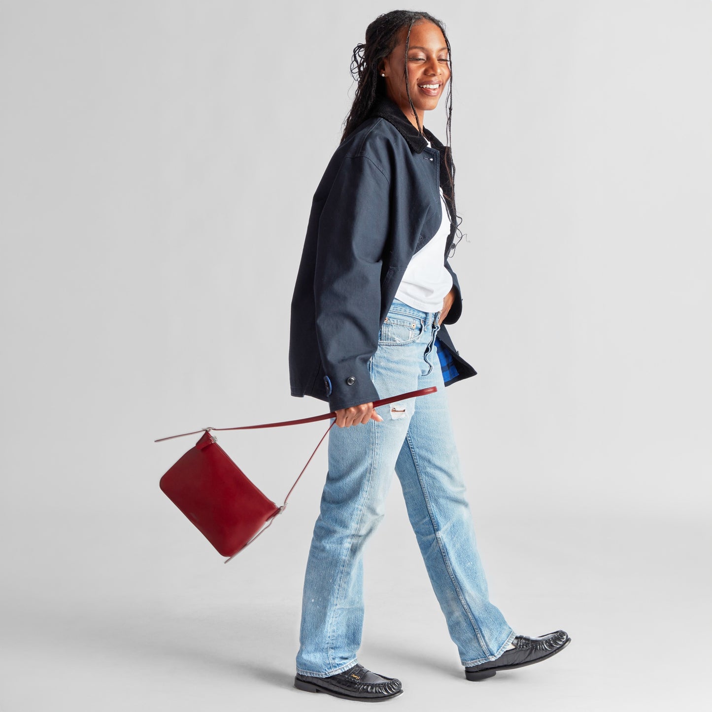 A smiling woman walks with a navy jacket, white shirt, light blue jeans, and black shoes, swinging her Shinola Small Bixby Crossbody - Ruby in Natural Leather by its strap against a plain light background.