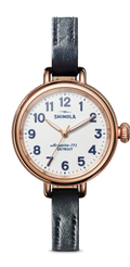 The Shinola Birdy Watch - White features a 34mm rose gold PVD case, round white dial with blue hour markers and gold hands, and a navy leather strap. The face reads "Shinola" and "Argonite-773 Detroit.