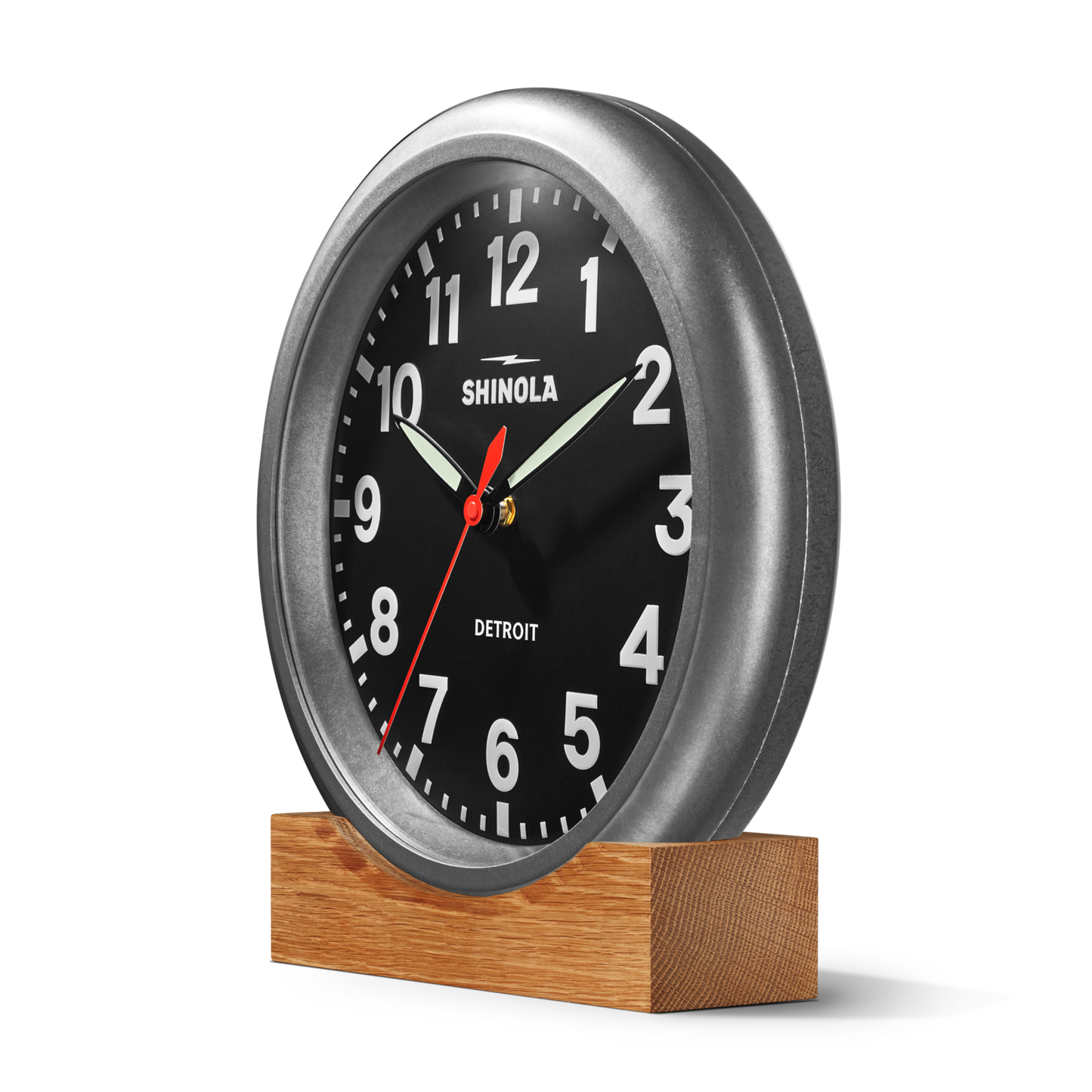 The Shinola 14" Runwell Clock in tumbled zinc features a black face with white numbers and hands, a red second hand, and sits on a wooden base against a plain white background.