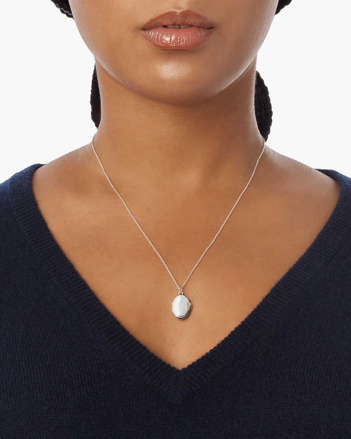 A woman wears a deep navy V-neck sweater and the Shinola Heirloom Petite Locket Necklace in sterling silver, featuring an oval pendant, shown from shoulders to just above the lips against a plain background.