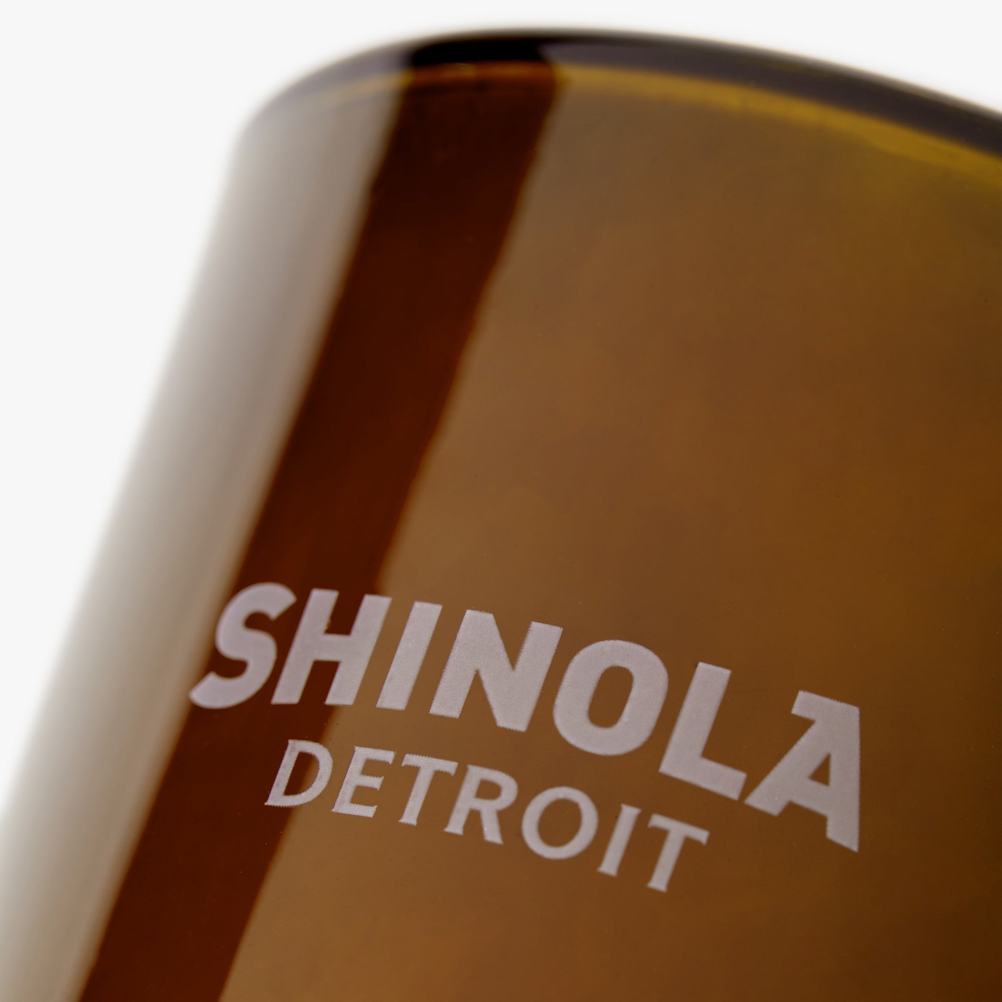 Close-up of a brown glass Shinola Smoke 8oz Scented Candle, with "Shinola Detroit" in white letters on the side, evoking a cozy autumn with its woody aroma.