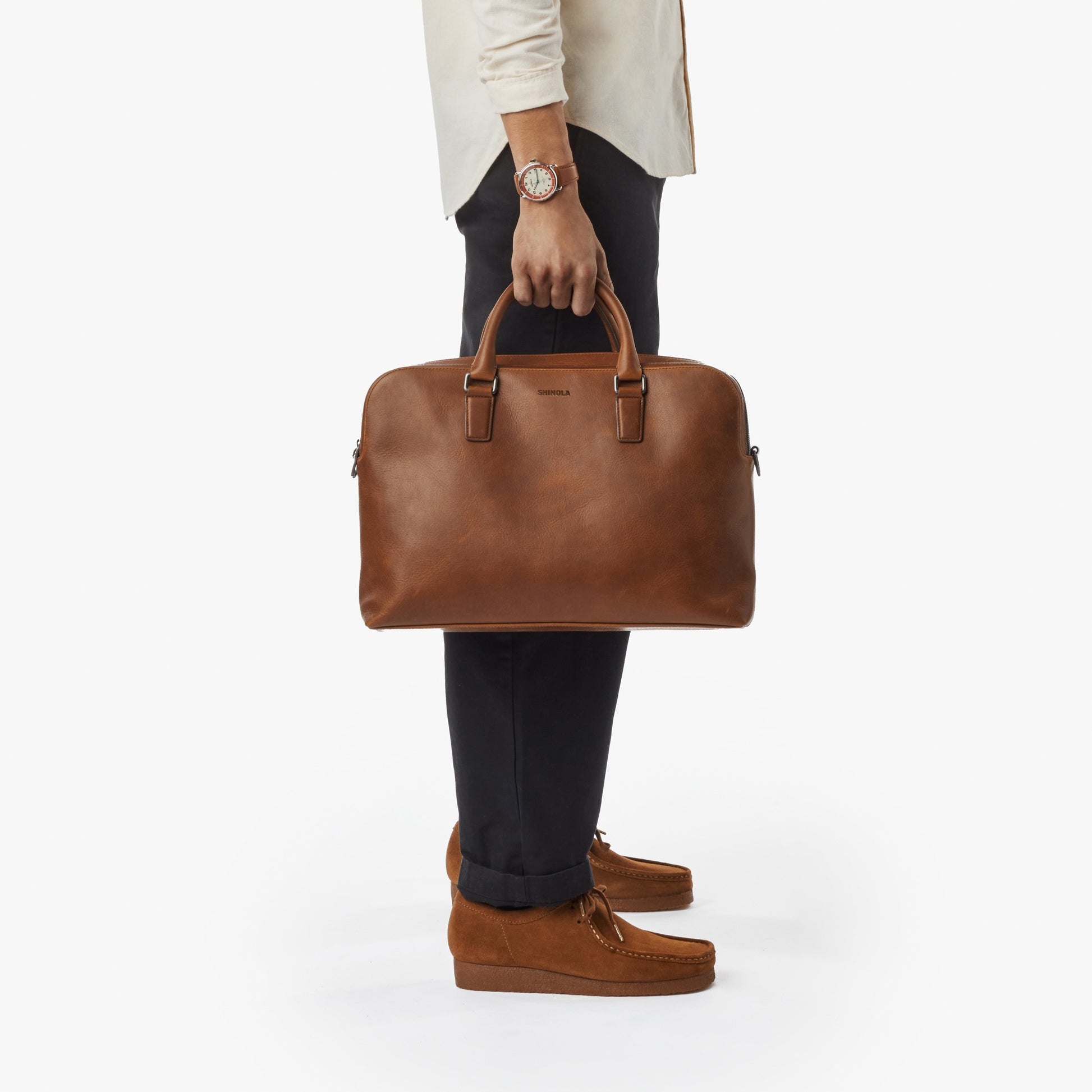 A person in black pants, tan shoes, and a white shirt holds the Shinola Double Zip Briefcase - Medium Brown by its handles against a plain white background.