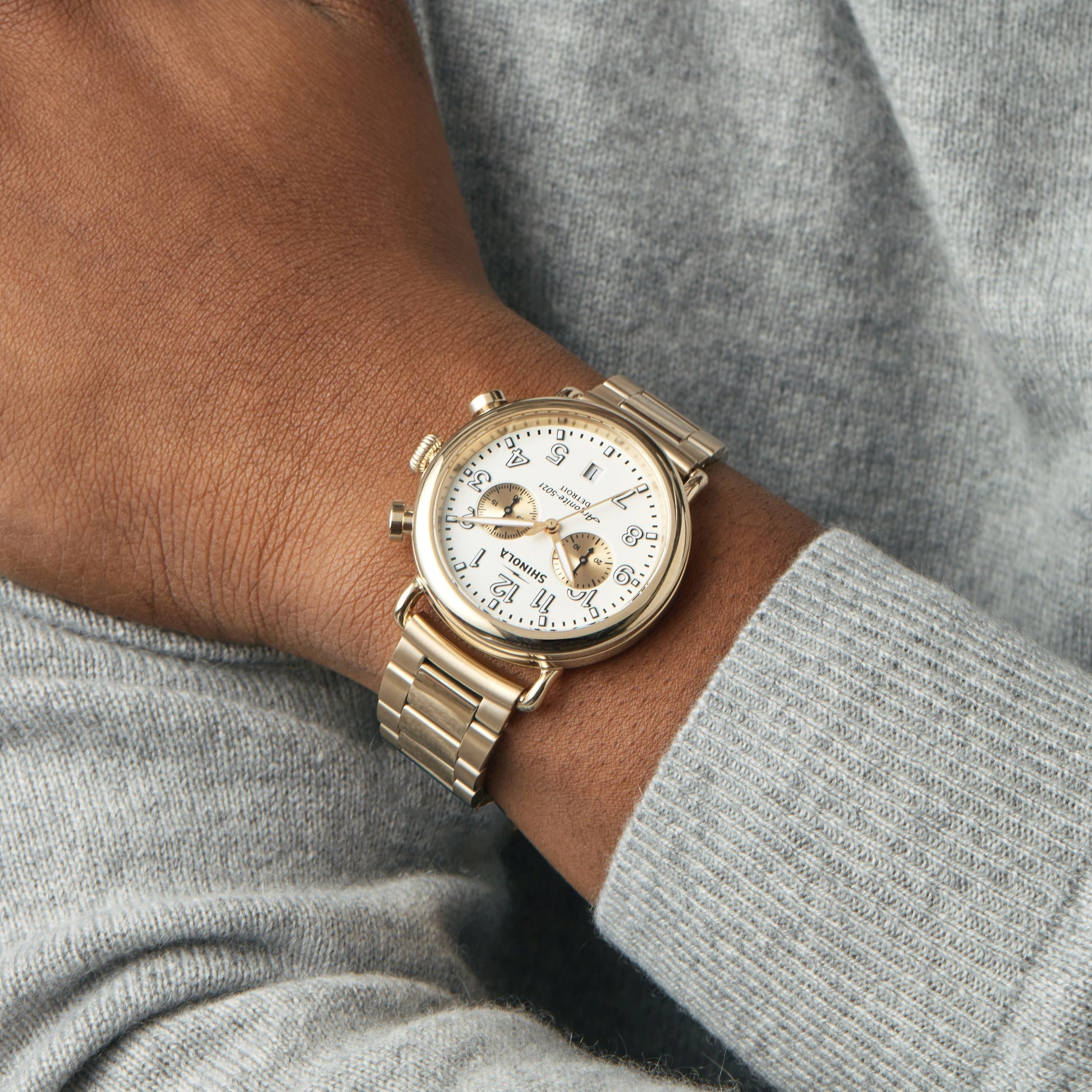 A person in a light gray sweater wears the Shinola Runwell Chronograph Watch - Ivory, featuring a gold stainless steel bracelet, white dial with multiple dials, displayed on their left wrist.