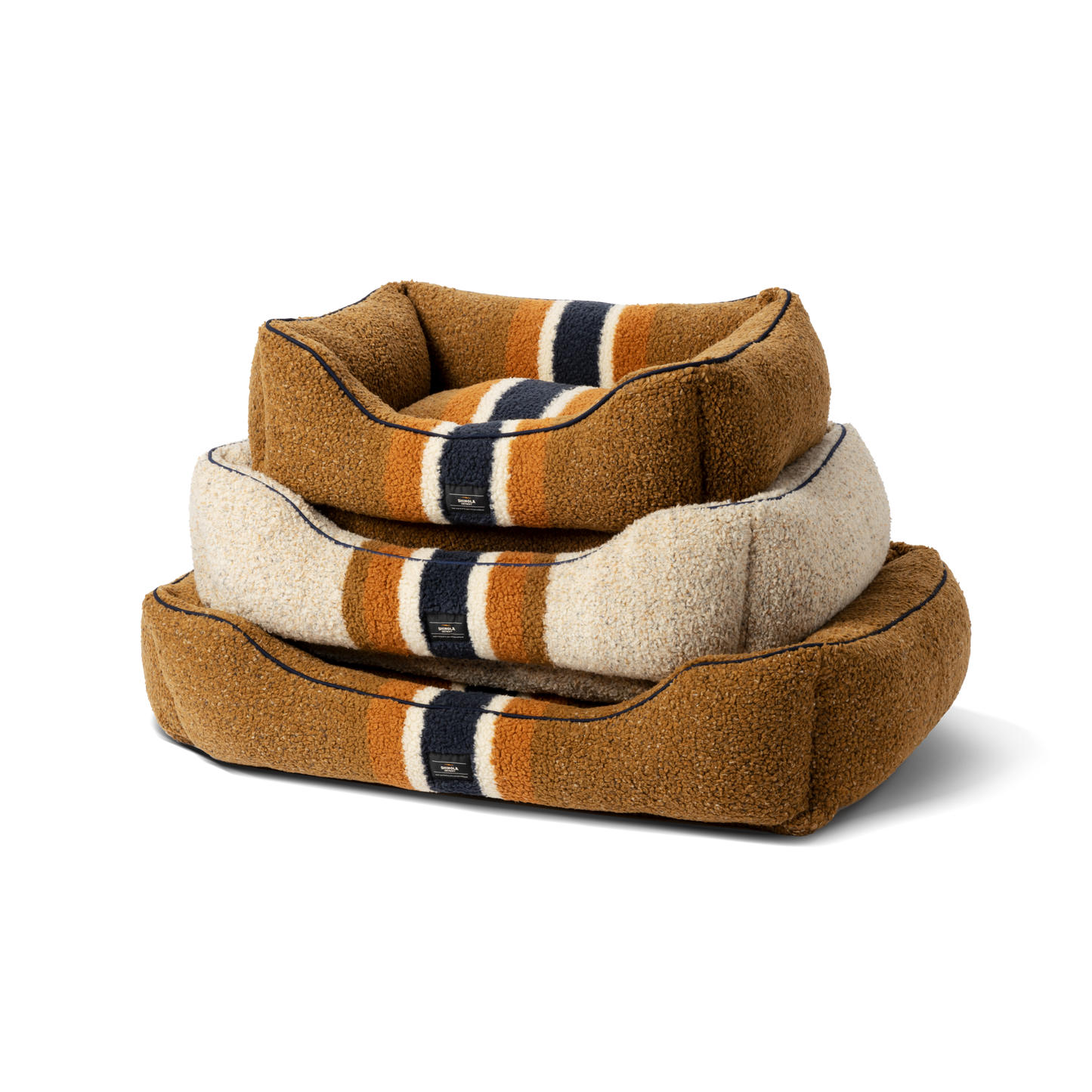 Three Shinola Kuddle Dog Beds in various sizes are stacked. Each Stripe Brown bed has memory fiber, raised edges, brown with cream, orange, and navy stripes, and is machine washable. The photo is on a plain white background.
