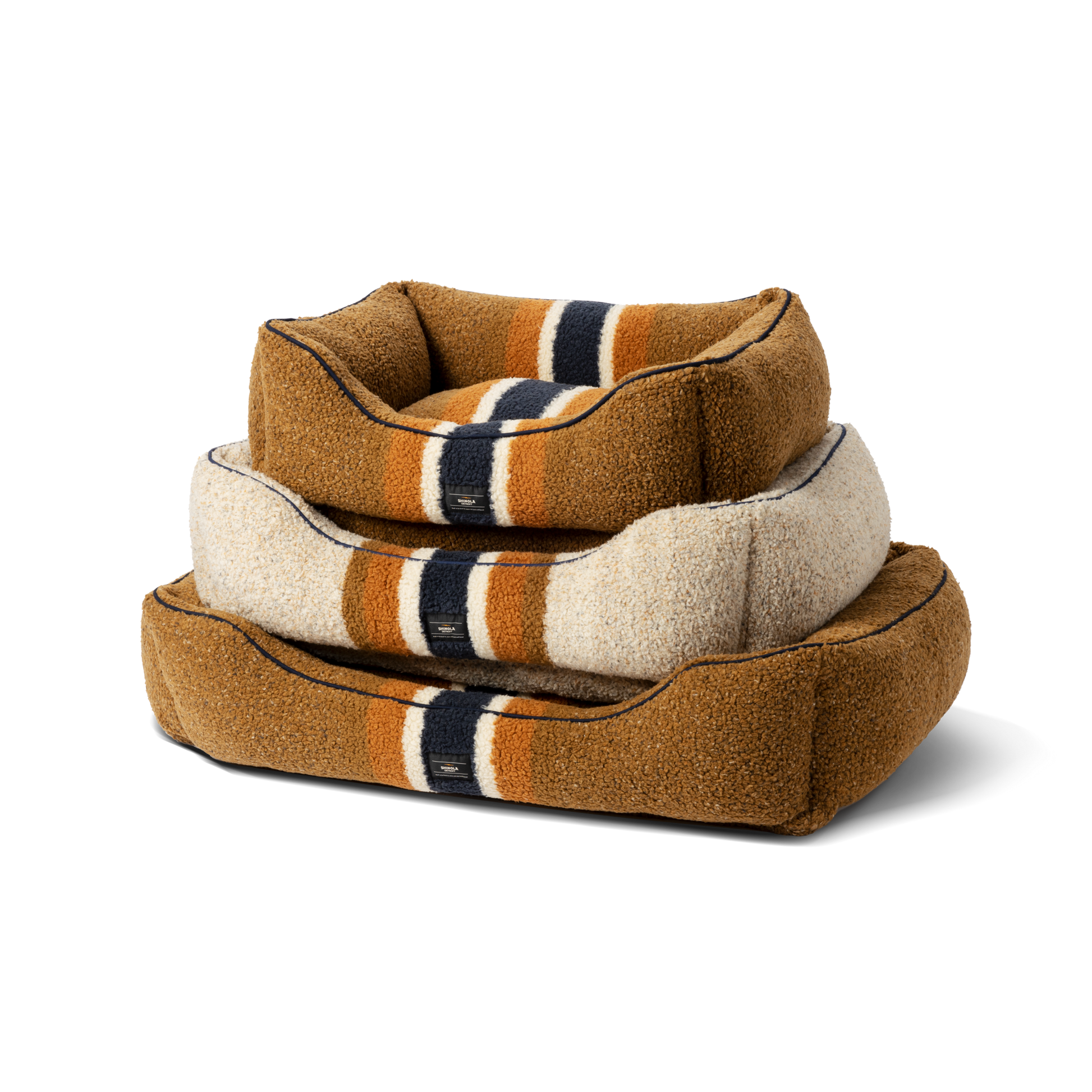 Three Shinola Kuddle Dog Beds in various sizes are stacked. Each Stripe Brown bed has memory fiber, raised edges, brown with cream, orange, and navy stripes, and is machine washable. The photo is on a plain white background.