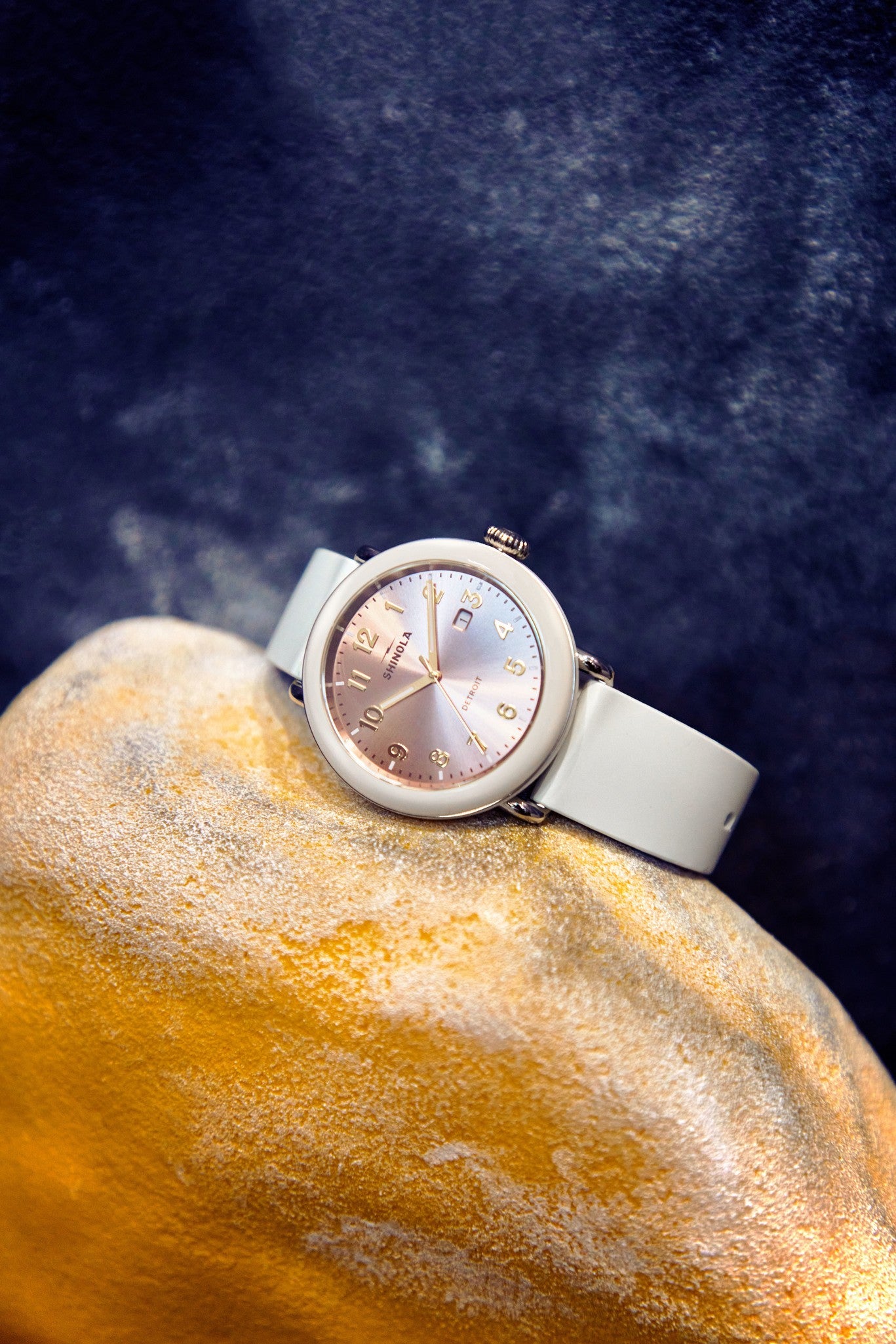 The Shinola Detrola Watch - Nude, featuring a light band and round iridescent face with gold numbers, rests on a yellow-beige textured rock against a dark, blurred background—a nod to classic American design.