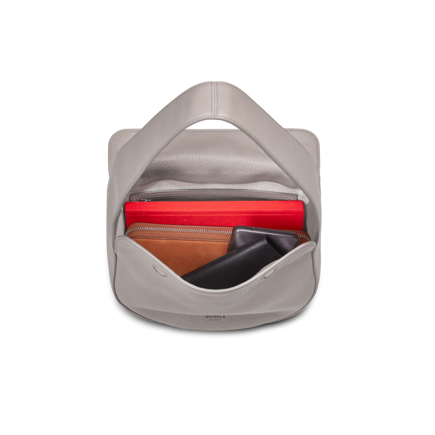 A top-down view of an open Shinola Small Shoulder Bag in taupe, neatly holding a red pouch, a brown wallet, and a black sunglasses case.