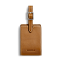 The Shinola Luggage Tag - Tan is crafted from Italian leather with white stitching, a silver buckle, and SHINOLA embossed at the bottom. Personalize it with monogramming. Shown against a plain white background.